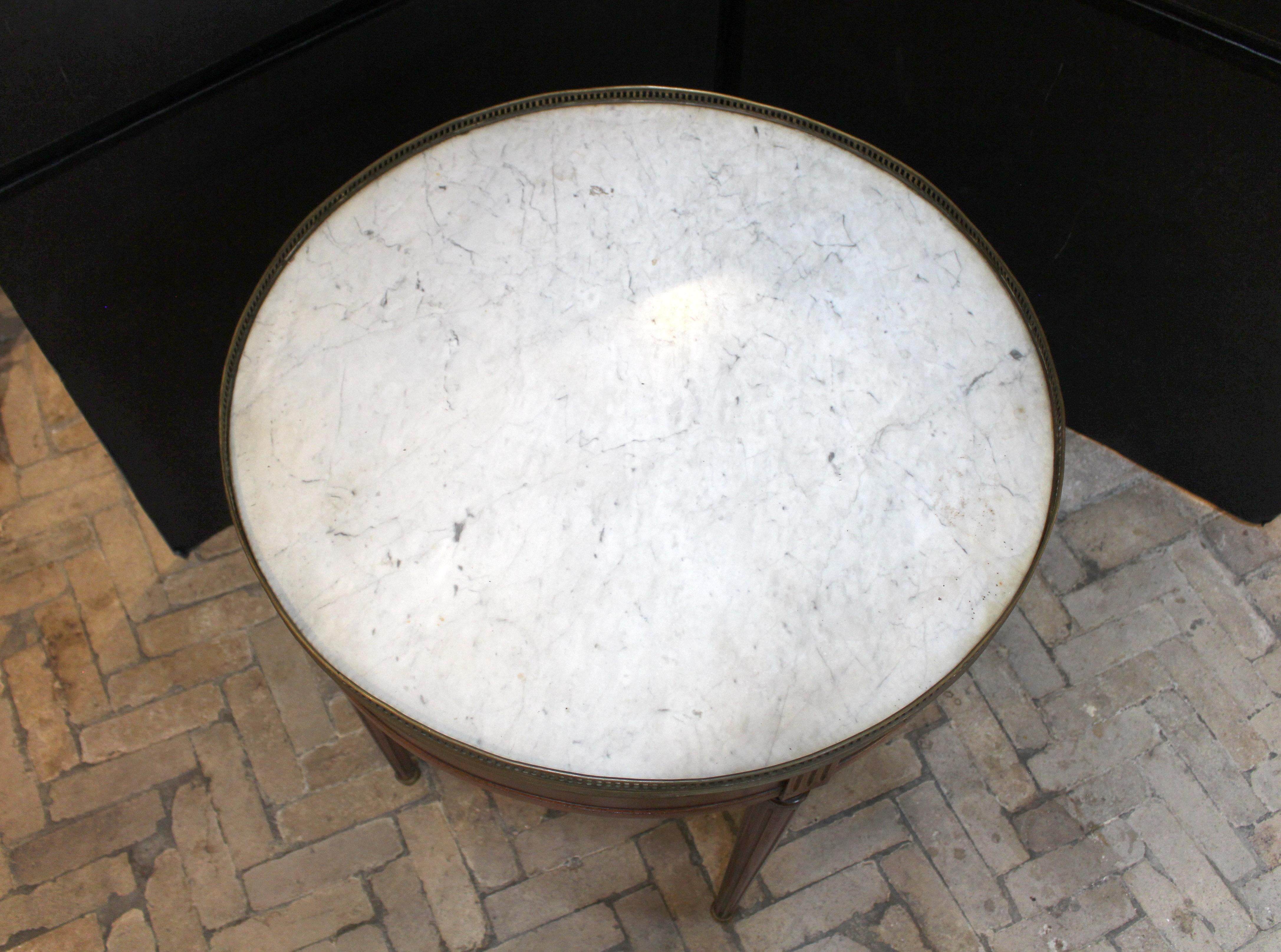 Mesa bouillotte con tapa de mármol de finales del siglo XIX, estilo Luis XVI, francesa. Caoba, encimera de mármol blanco con estrías grises. Galería y sabots de latón. Patas acanaladas, torneadas y ahusadas, rematadas por un friso acanalado. Cajones