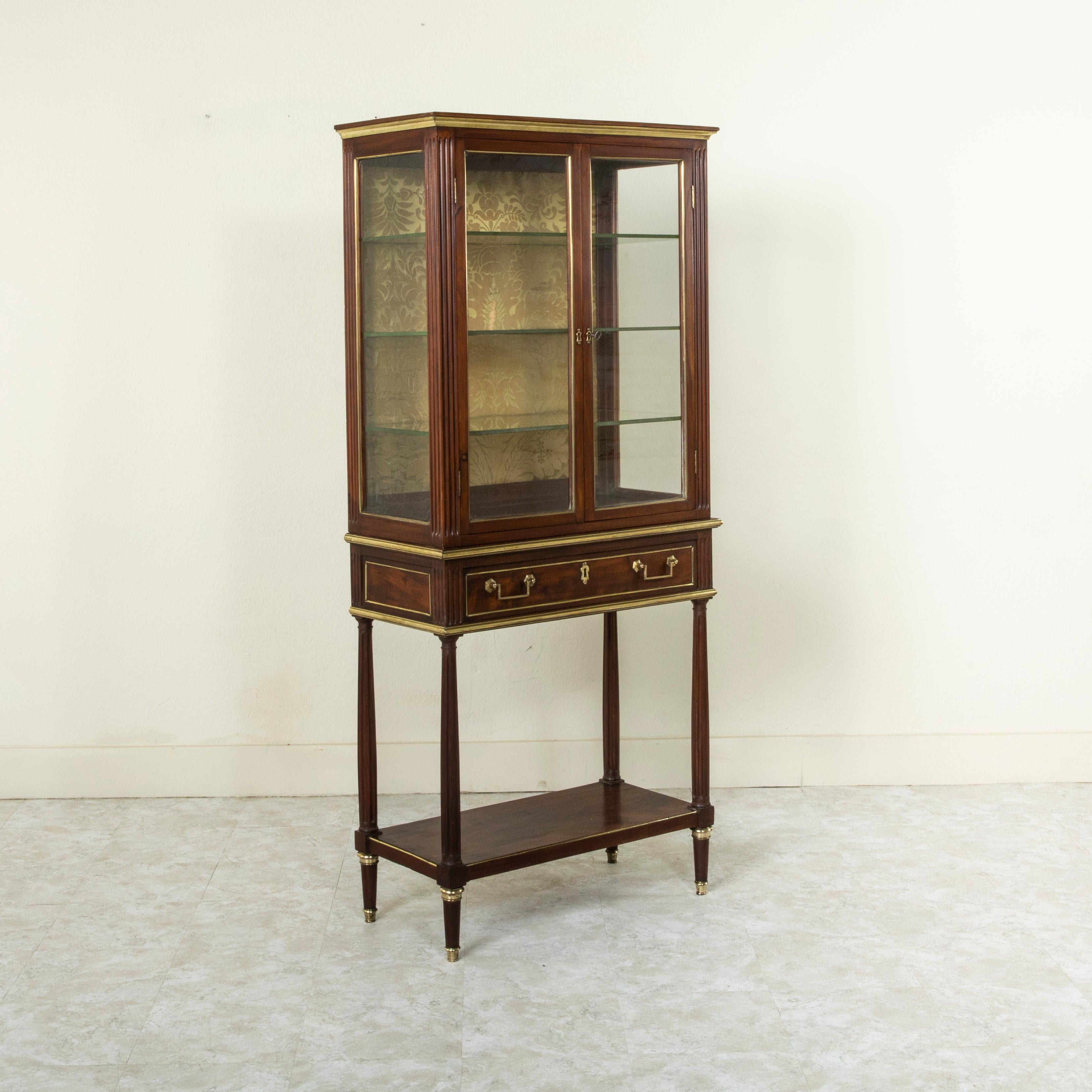 This mid nineteenth century French Louis XVI style mahogany console vitrine features fluted corners and bronze banding on its facade and sides. The upper cabinet is finished with glass on three sides and its two doors open to access its fabric lined