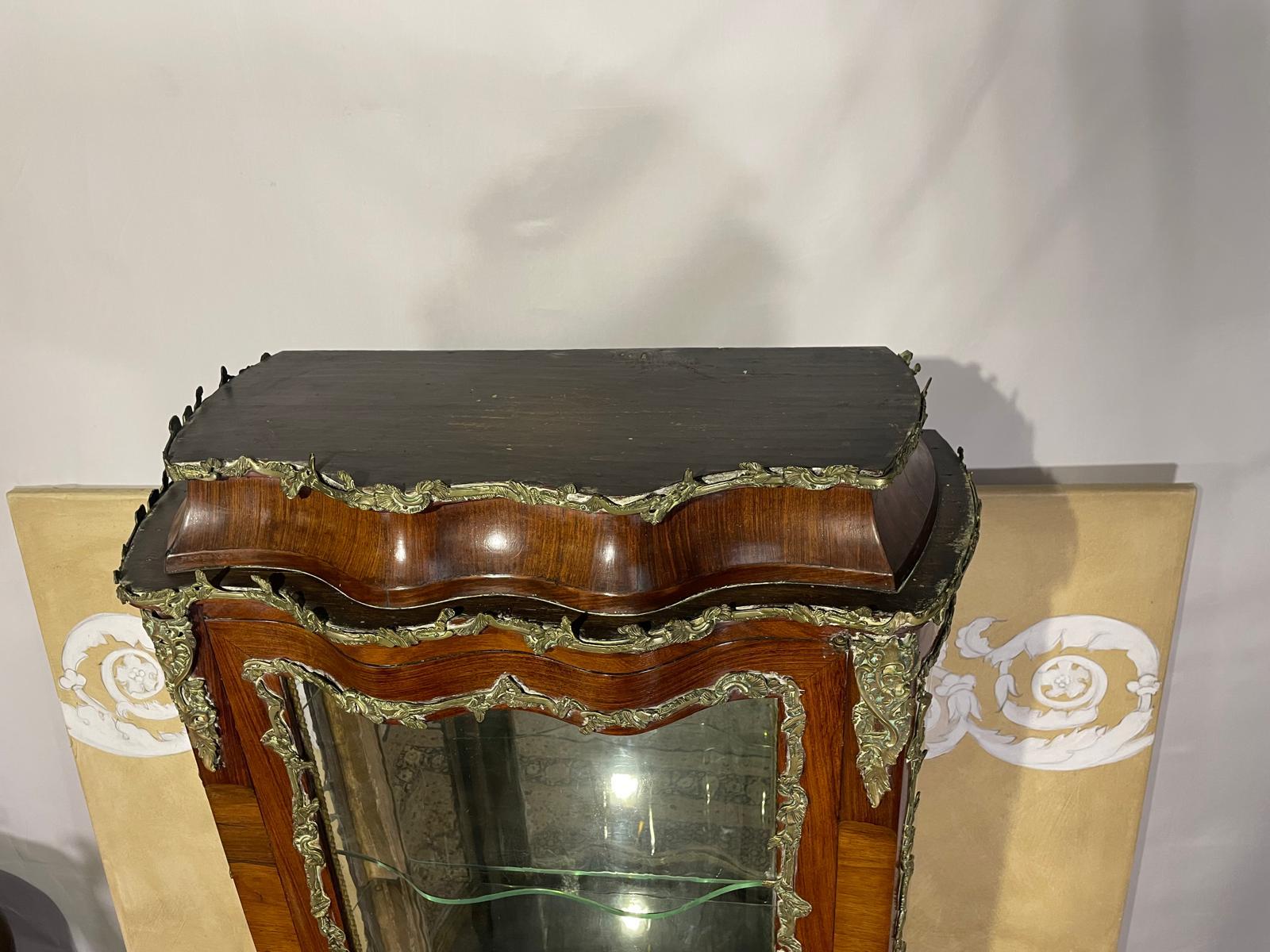 Armadio vitreo in stile Luigi XVI, risalente alla fine del XIX secolo, caratterizzato da un'elegante forma a serpentina con un'anta in vetro smussato e lati in vetro curvo. Il mobile è riccamente decorato con ornamenti in bronzo dorato e ormolu,