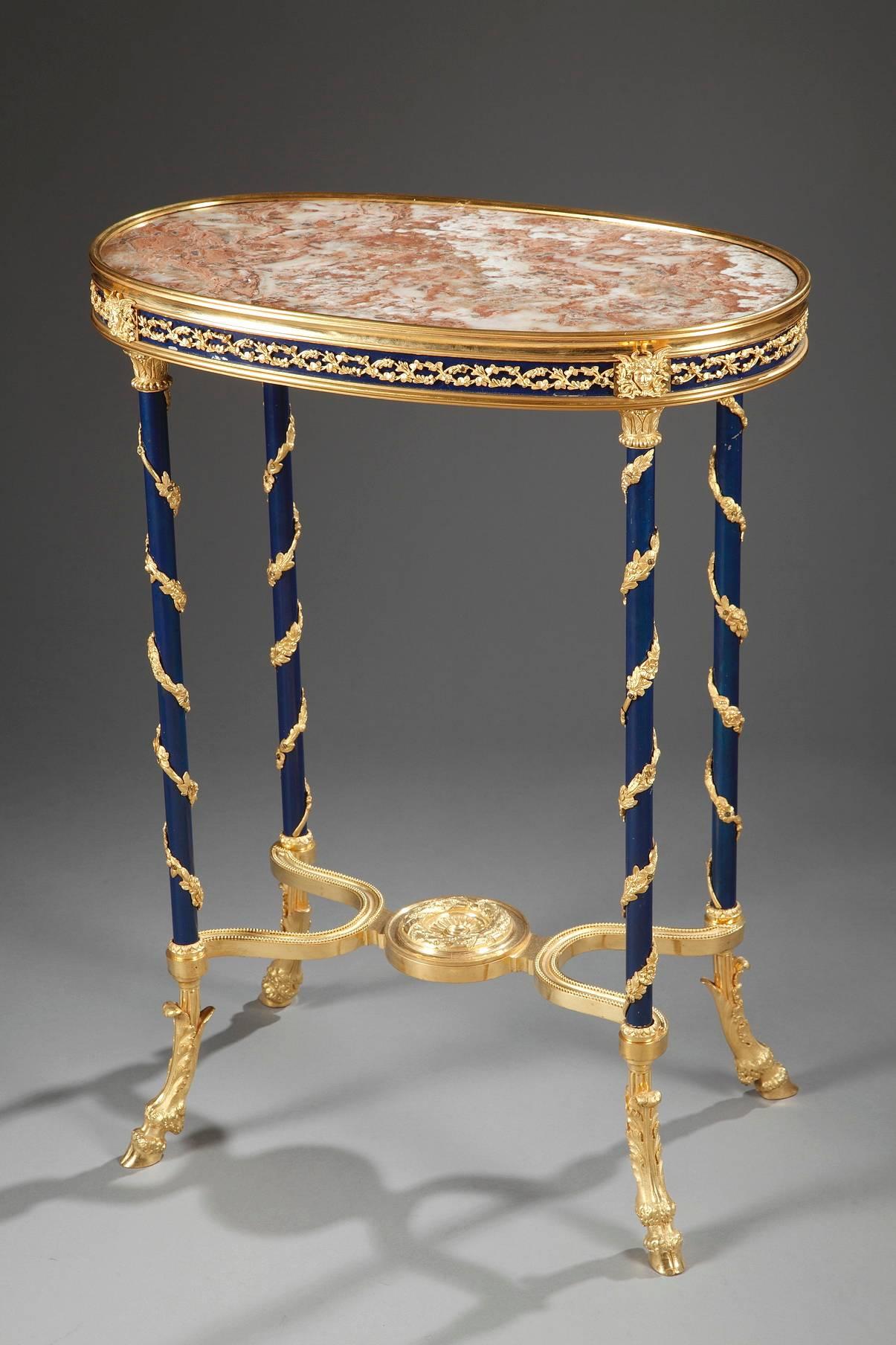 Gueridon with its oval, marble tray. The marble top is framed with an ormolu frieze of interlacing foliage set on a dark blue background. Masks interrupt the frieze above the four legs of the table, which are slender, dark-blue columns. The tops of