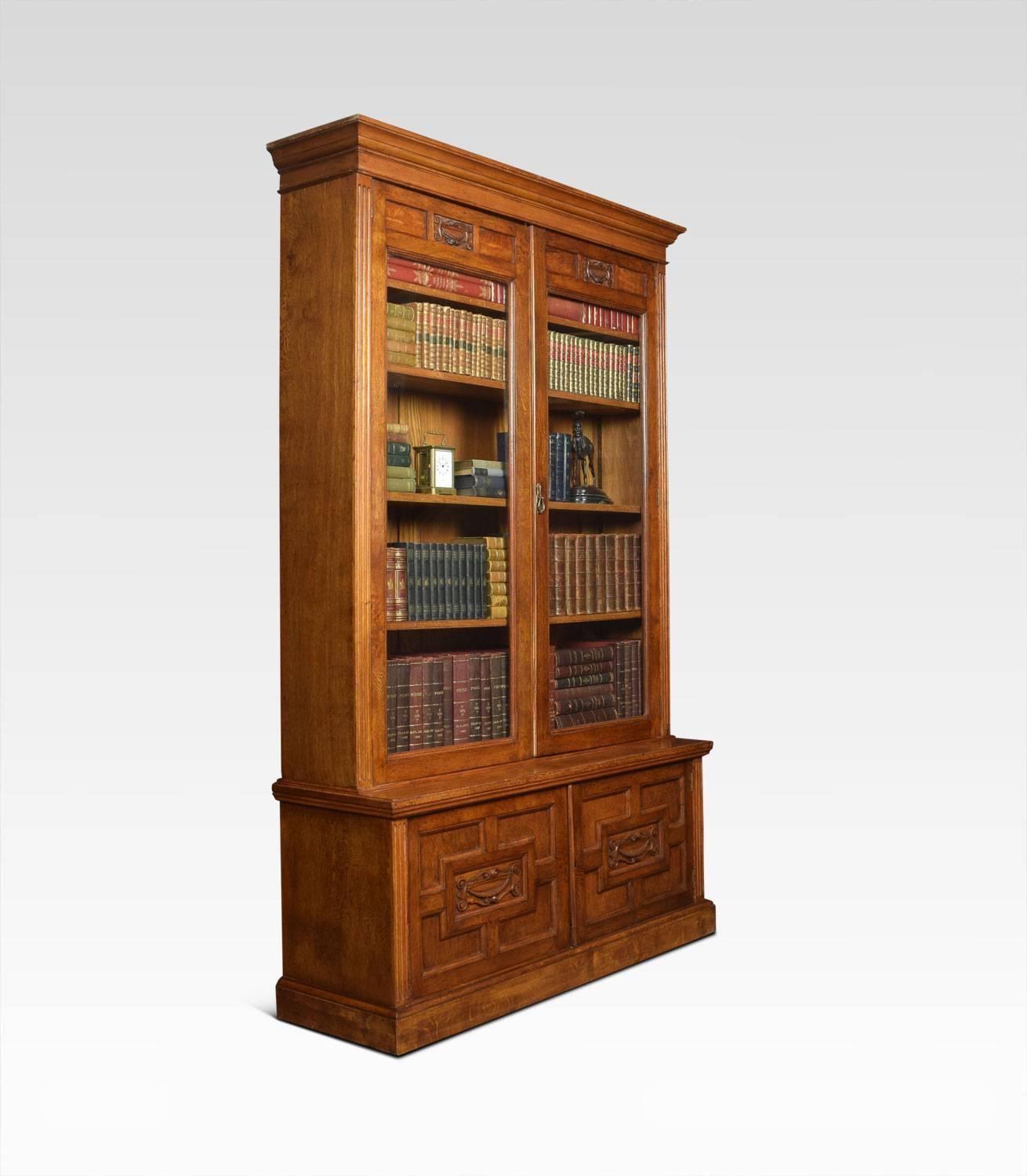 Solid oak two-door bookcase, the projecting cornice, above carved freeze leading down two a large pair of glazed doors. Opening to reveal four adjustable shelves. The base fitted with two geometric carved paneled doors opening to reveal single fixed