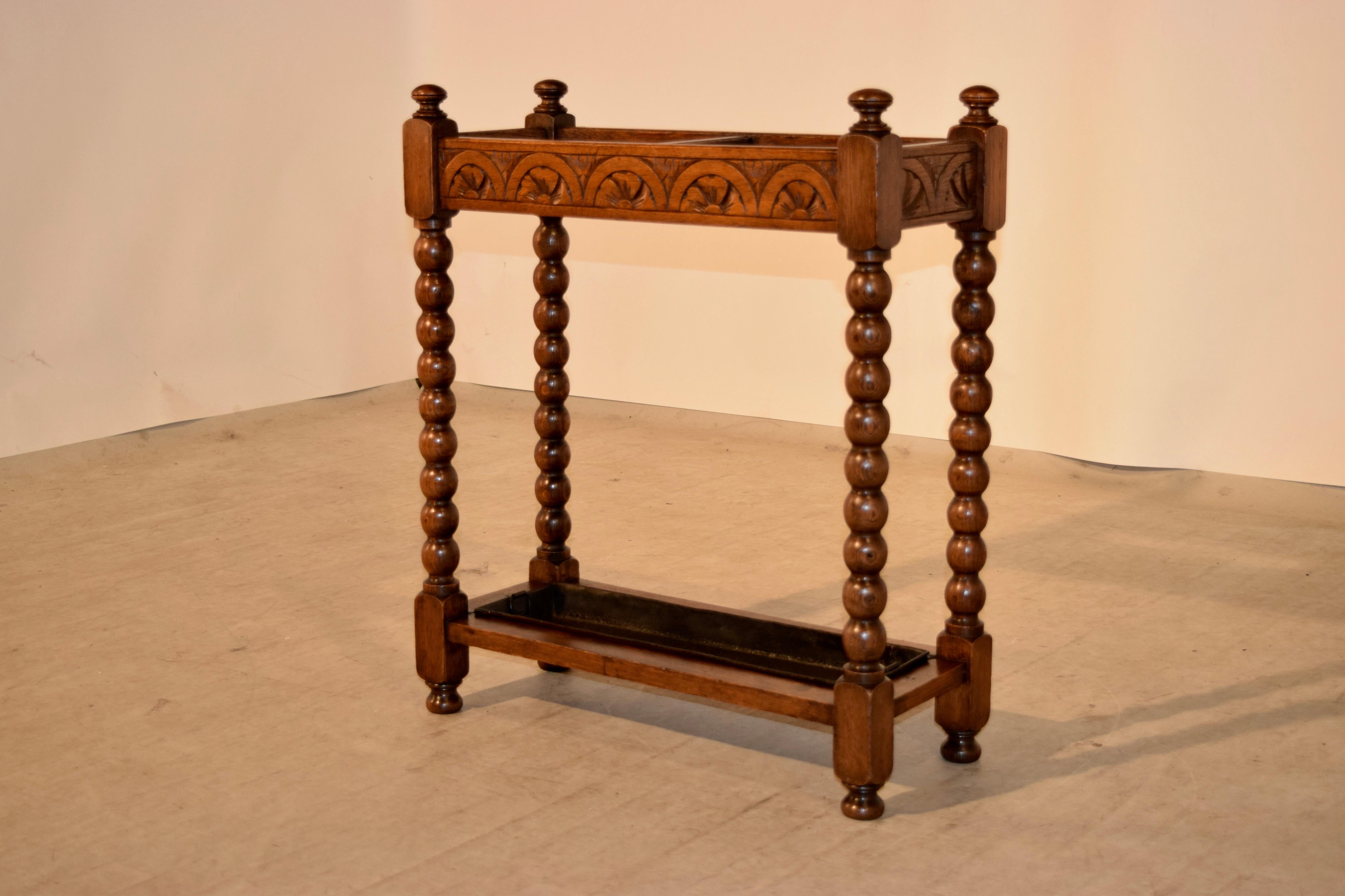Late 19th century oak umbrella stand from England with hand-carved decorated top rail, joined by hand-turned spool legs, which are joined at the bottom by simple stretchers with a metal drip tray. Supported on hand-turned feet.