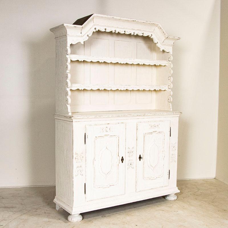 This charming antique oak cupboard was given a (newer) white painted finish that has been slightly distressed to compliment the European country style of the display cabinet. The scalloped edging, bonnet/crown and hand-carved details adds a touch of