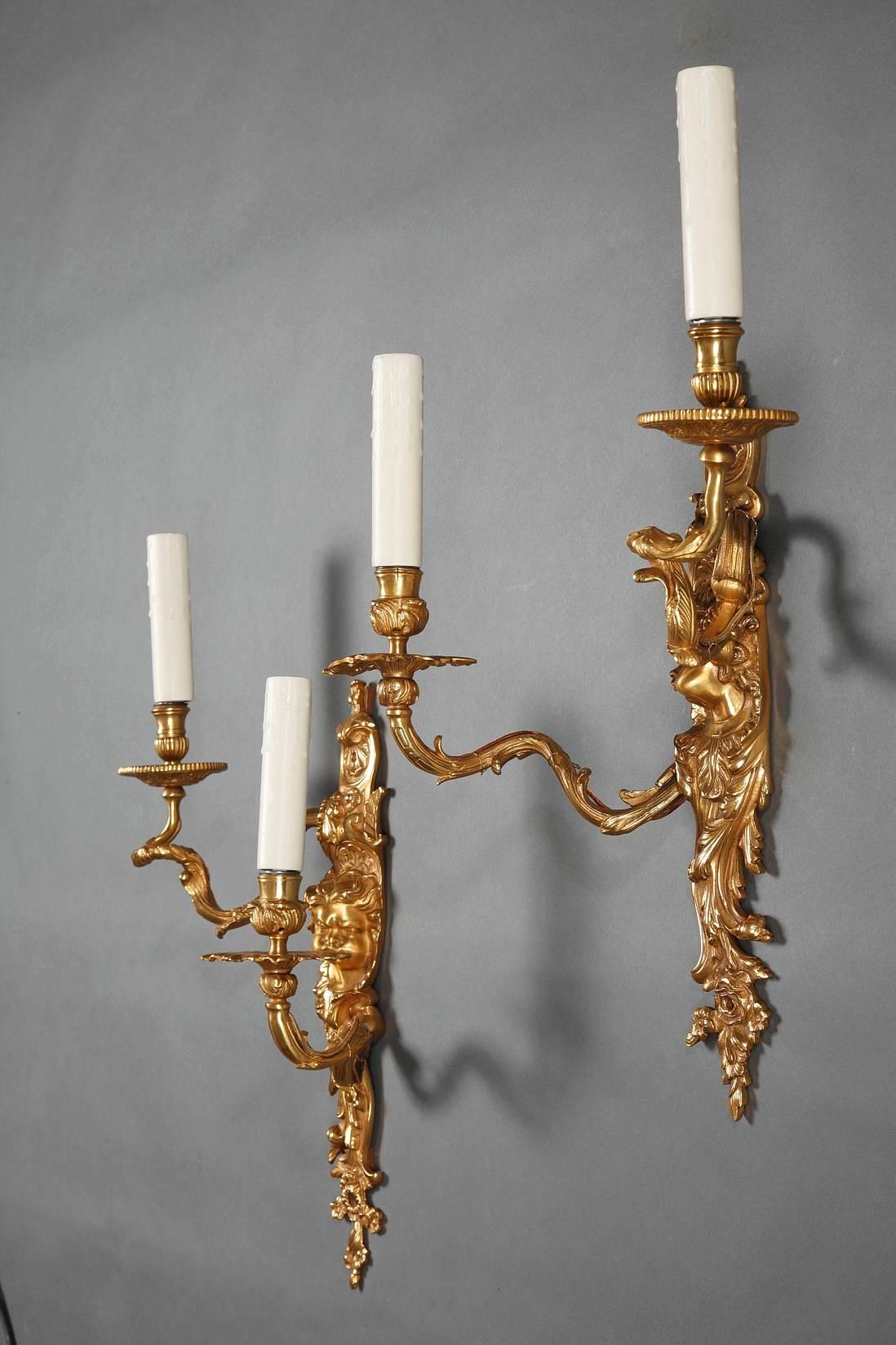 Large pair of gilded and sculpted bronze sconces in Louis XV style. They each feature two arms that wind out from the stem and upward like branches. Each stem is richly decorated with scrolling foliage, shells, flowers, and a baby who is puffing his