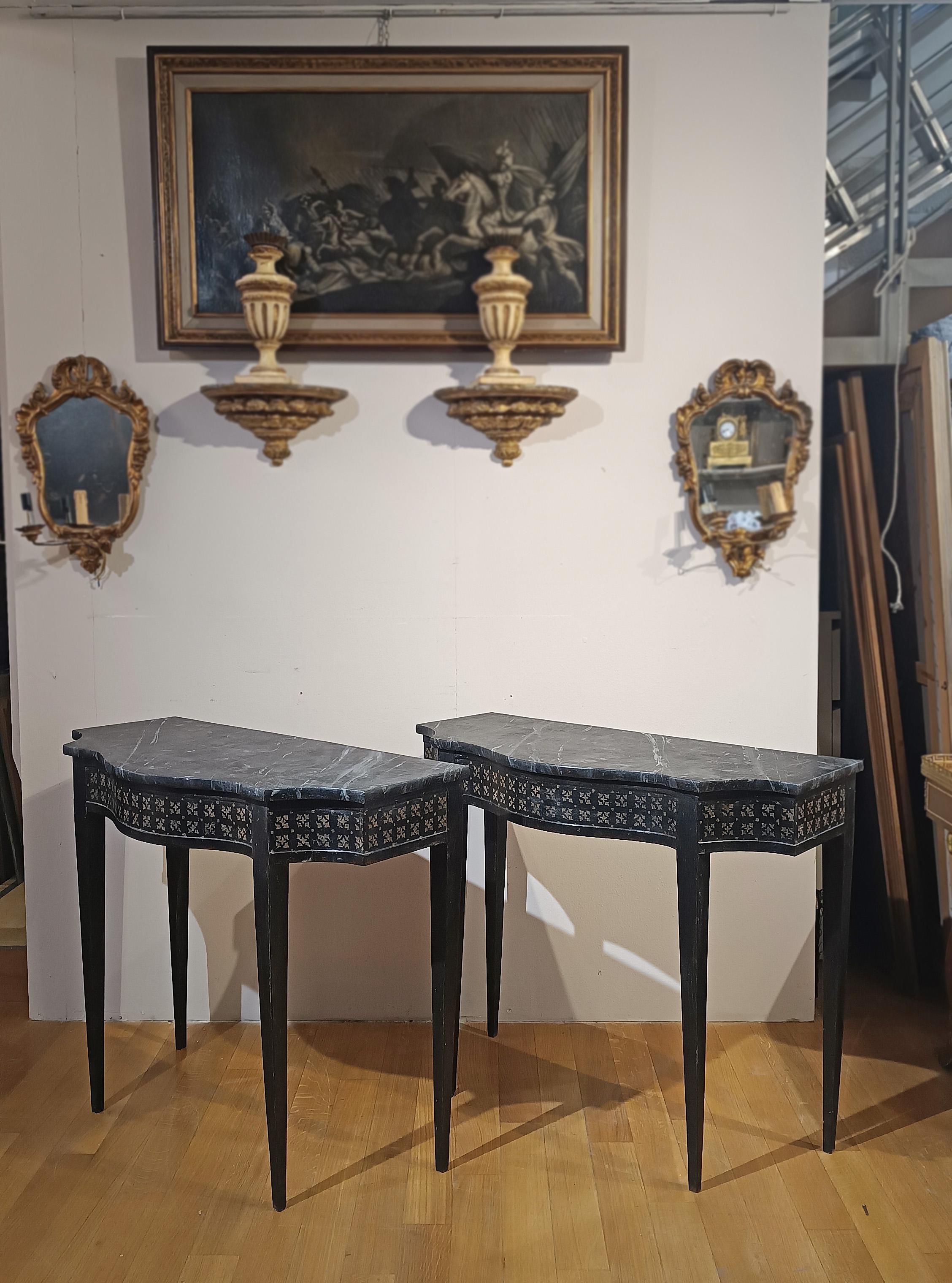 Elegant pair of carved wooden consolles tables, painted in two-tone black and white, with a chalky background. The consolles tables feature a curved top, truncated pyramid legs typical of the neoclassical style, and a painted faux marble top, which