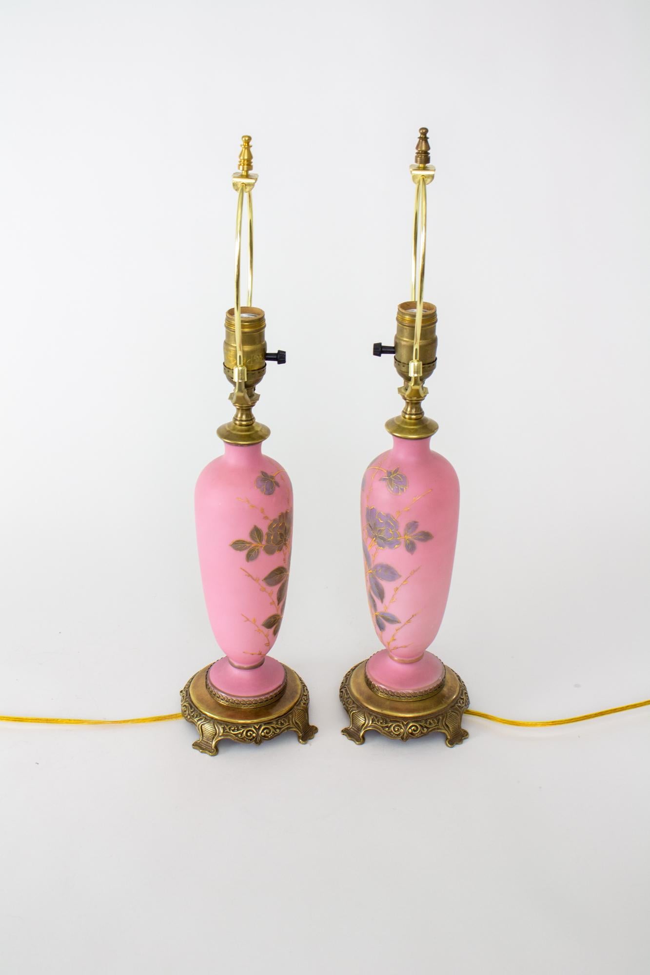Late 19th Century Austrian pink table lamps - a pair. These lamps are made of a pink glass in a vase form. They are decorated with purple painted flowers with raised gold detail. A dark cold painted band circles the stem and base. The lamps are