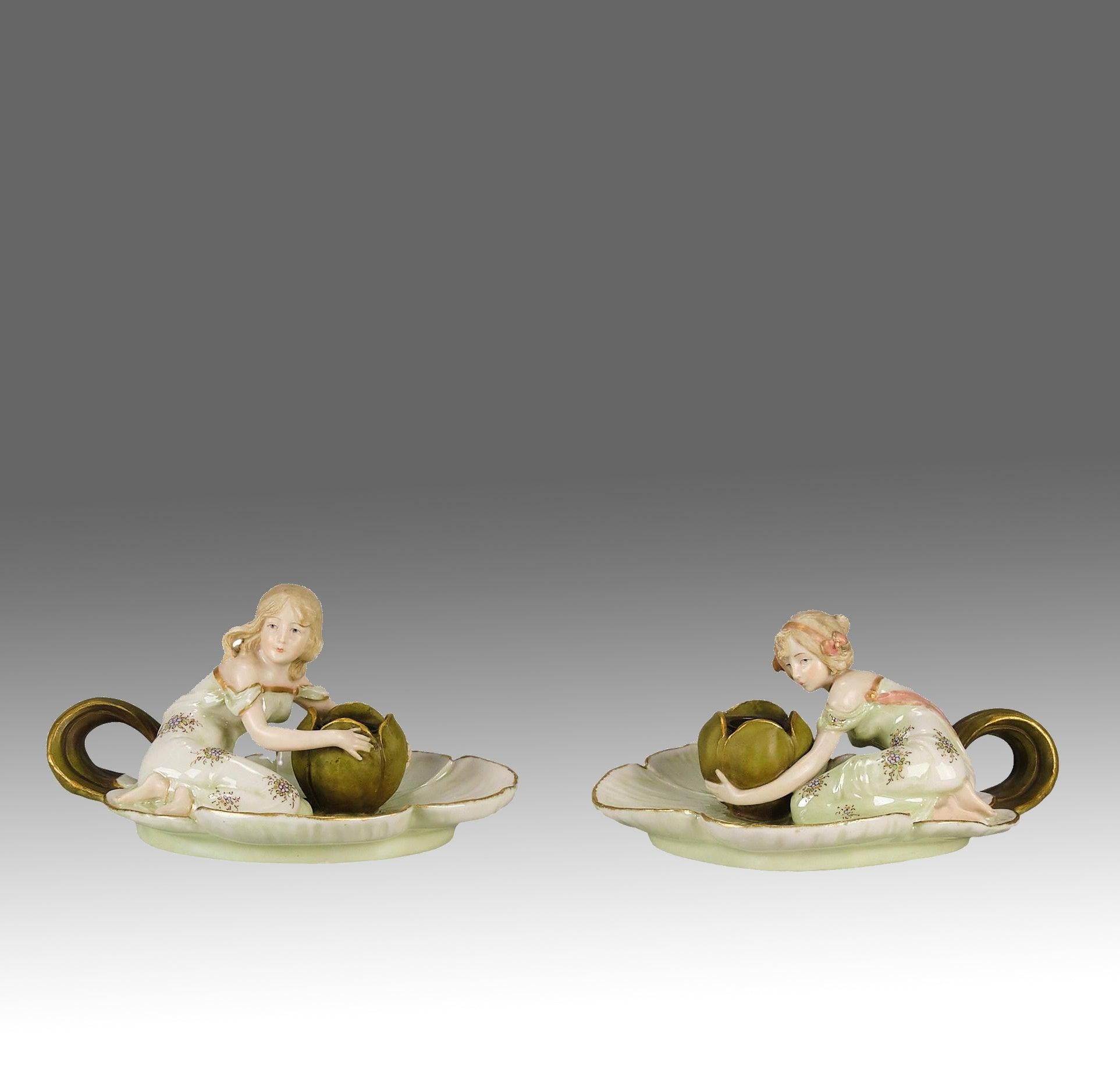 Atractiva pareja de candelabros de cerámica Art Nouveau en tenues tonos vitela con reflejos dorados, modelados con dos jovencitas arrodilladas dentro de platos de asas poco profundas con los brazos rodeando los candelabros que tienen forma de bulbos