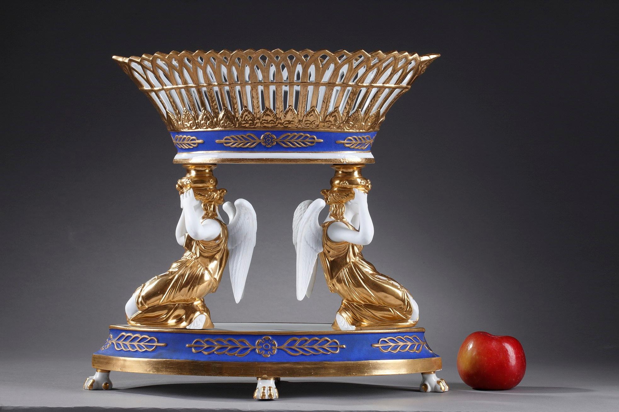 Late 19th century white, blue and gold hard-porcelain centerpiece featuring two winged women holding the pierced basket, highlighted with gold and palmette motif. The table centerpiece is set on an oblong base decorated with palmettes, above four