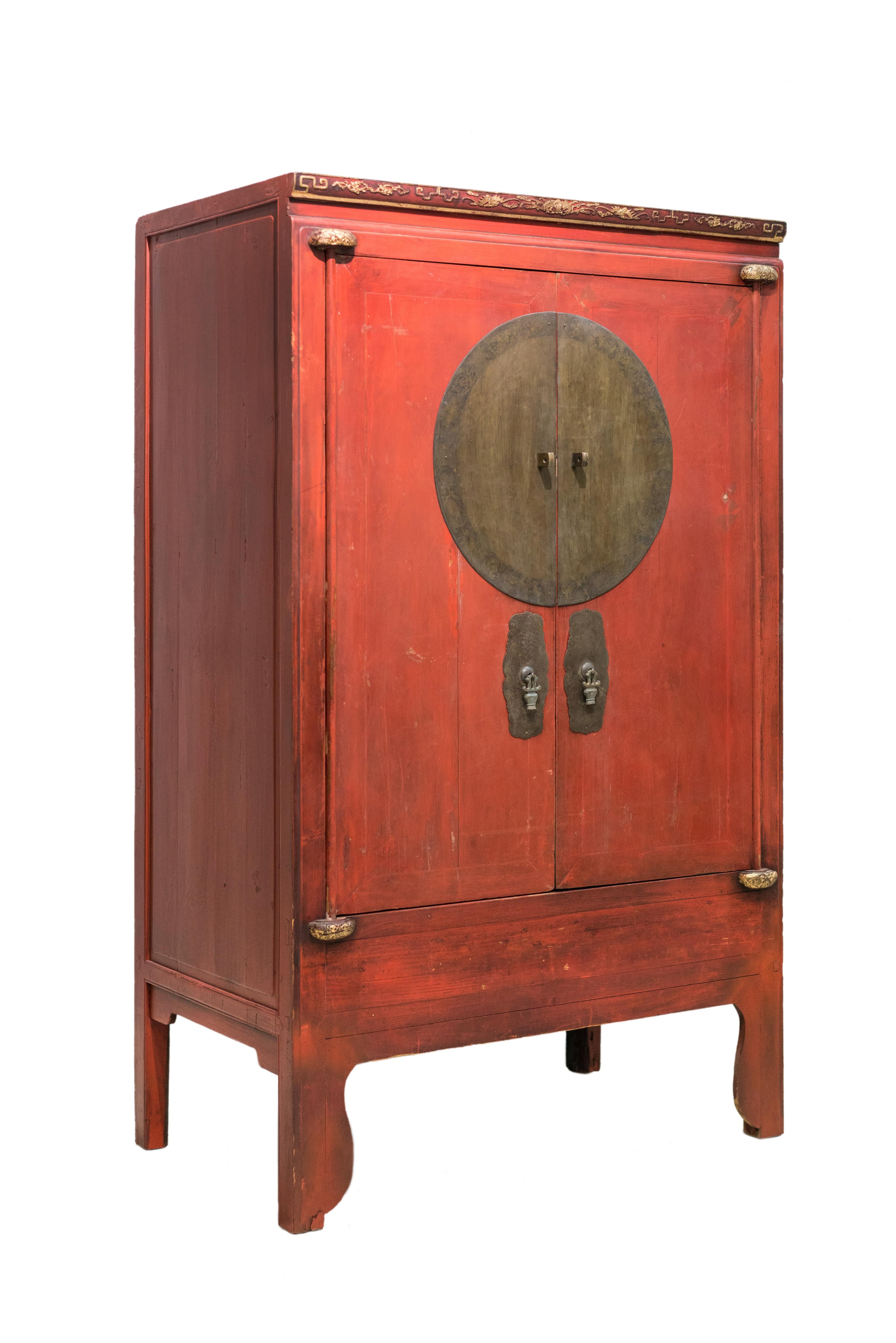 An antique red wedding cabinet from Zhejiang province. The carvings on the top front have designs of bats and clouds, symbolizing good fortune. The wooden knobs holding the hinges are intricately carved with a butterfly design. The round brass plate