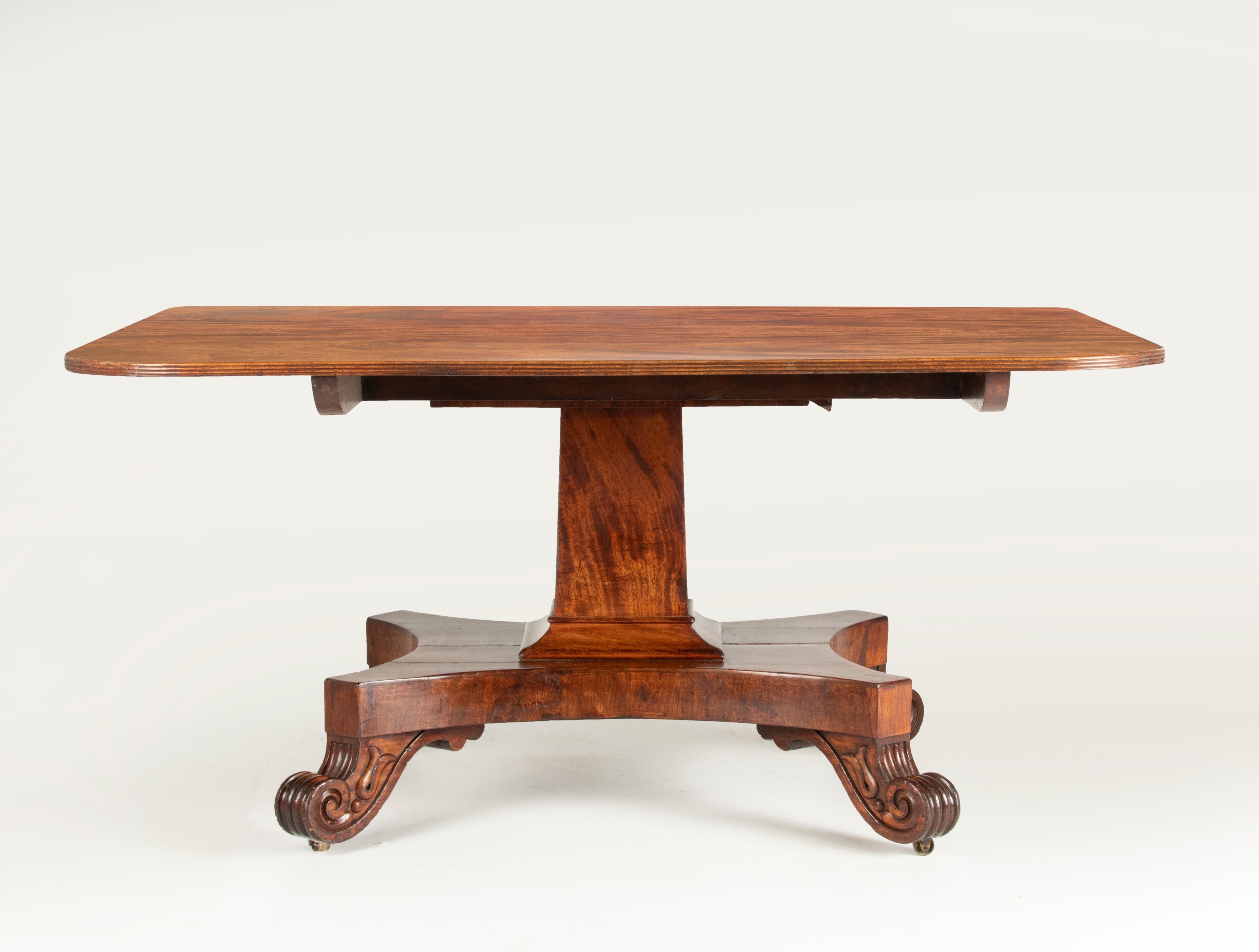 An Antique English Regency style dining room table with a rectangular top. The table is made of mahogany veneer, the top has a nice warm look and feel. Rests on a pedestal base with carved feet with small wheels underneath. Made in England, circa