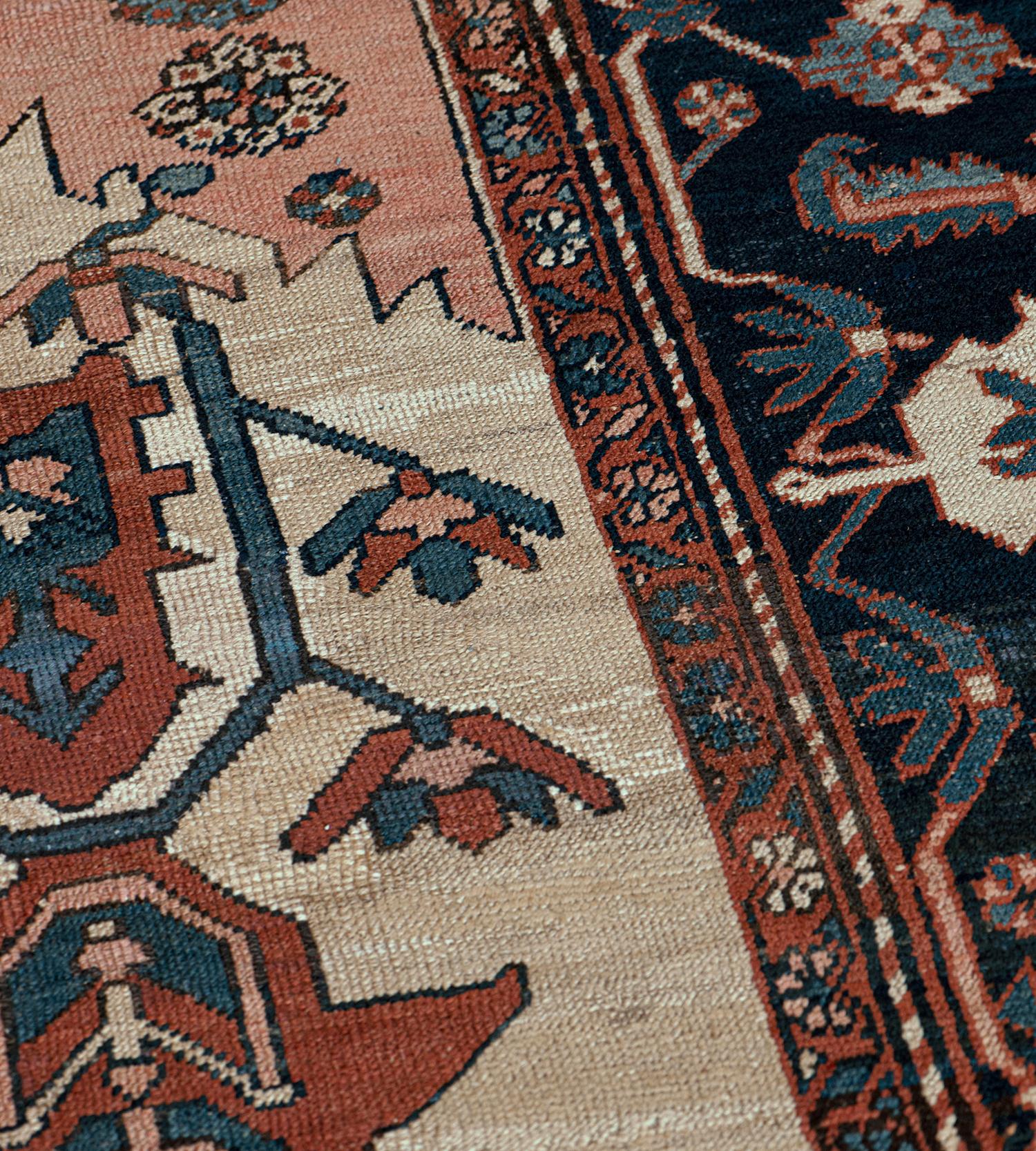 This traditional handwoven Persian Serapi rug has an ivory field with an overall design of brick-red, indigo and coral bold palmette and floral vines, in a deep indigo angular palmette vine border.