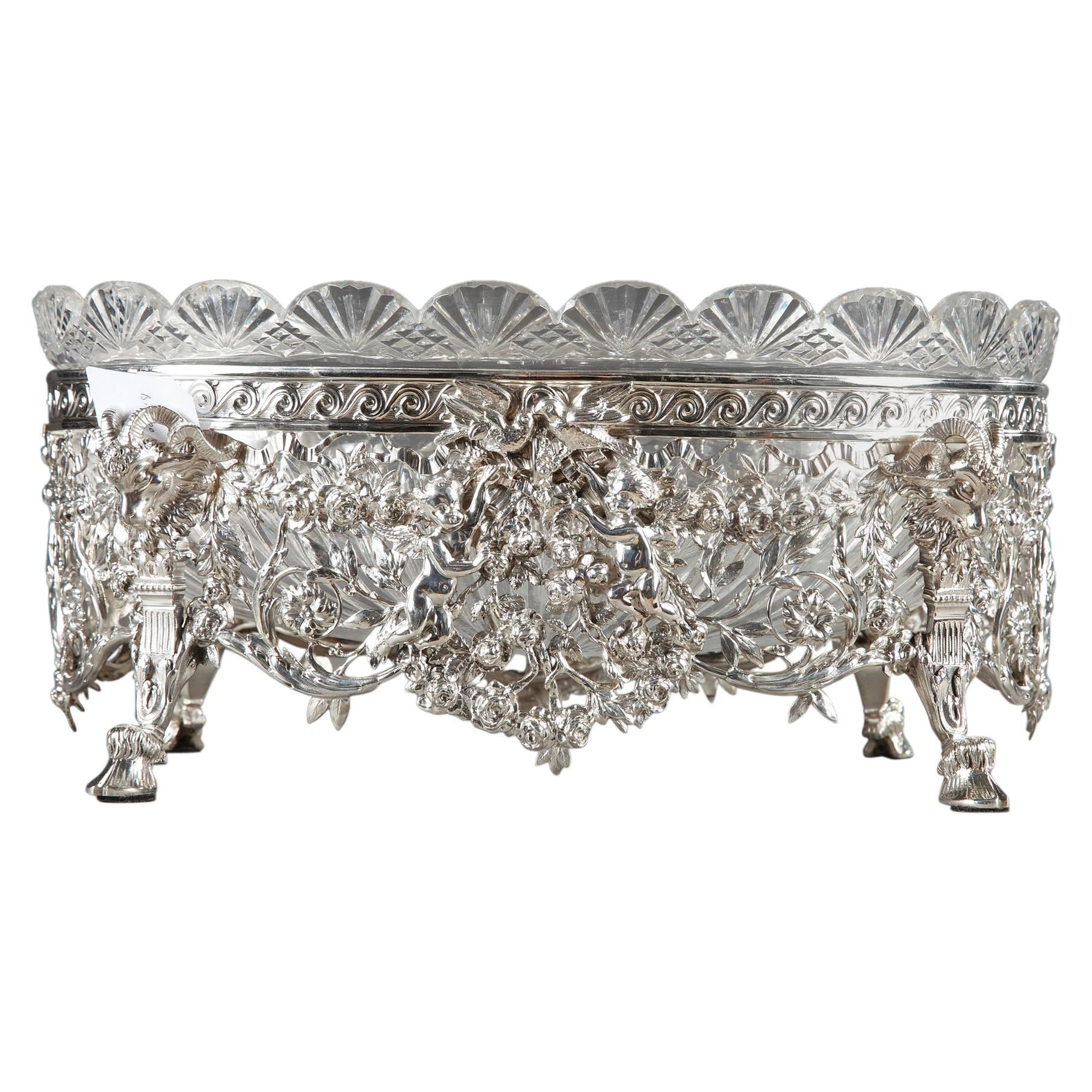 Silver jardinière composed of a cut-crystal, oval bowl resting in a silver frame. The scalloped edge of the crystal bowl is decorated with radiating and diamond-shaped patterns. The silver frame is richly decorated with openwork ribbons, Greek