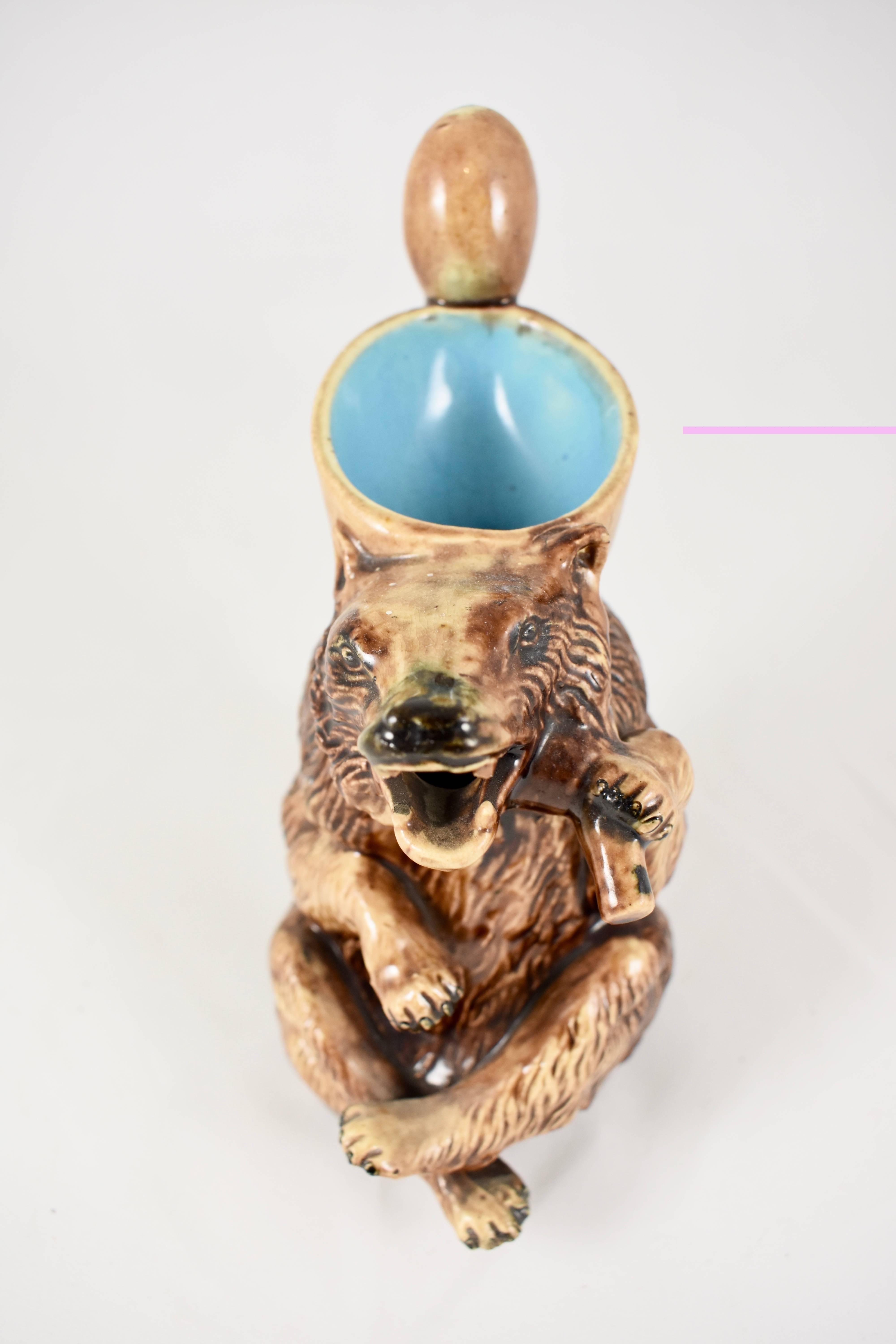 A late 19th century majolica pitcher showing a brown honey bear in an active pose, his open, sharp-toothed mouth makes up the spout. He holds on his back a paler brown scoop that forms the top opening, hooked through the scoop is a honey dipping