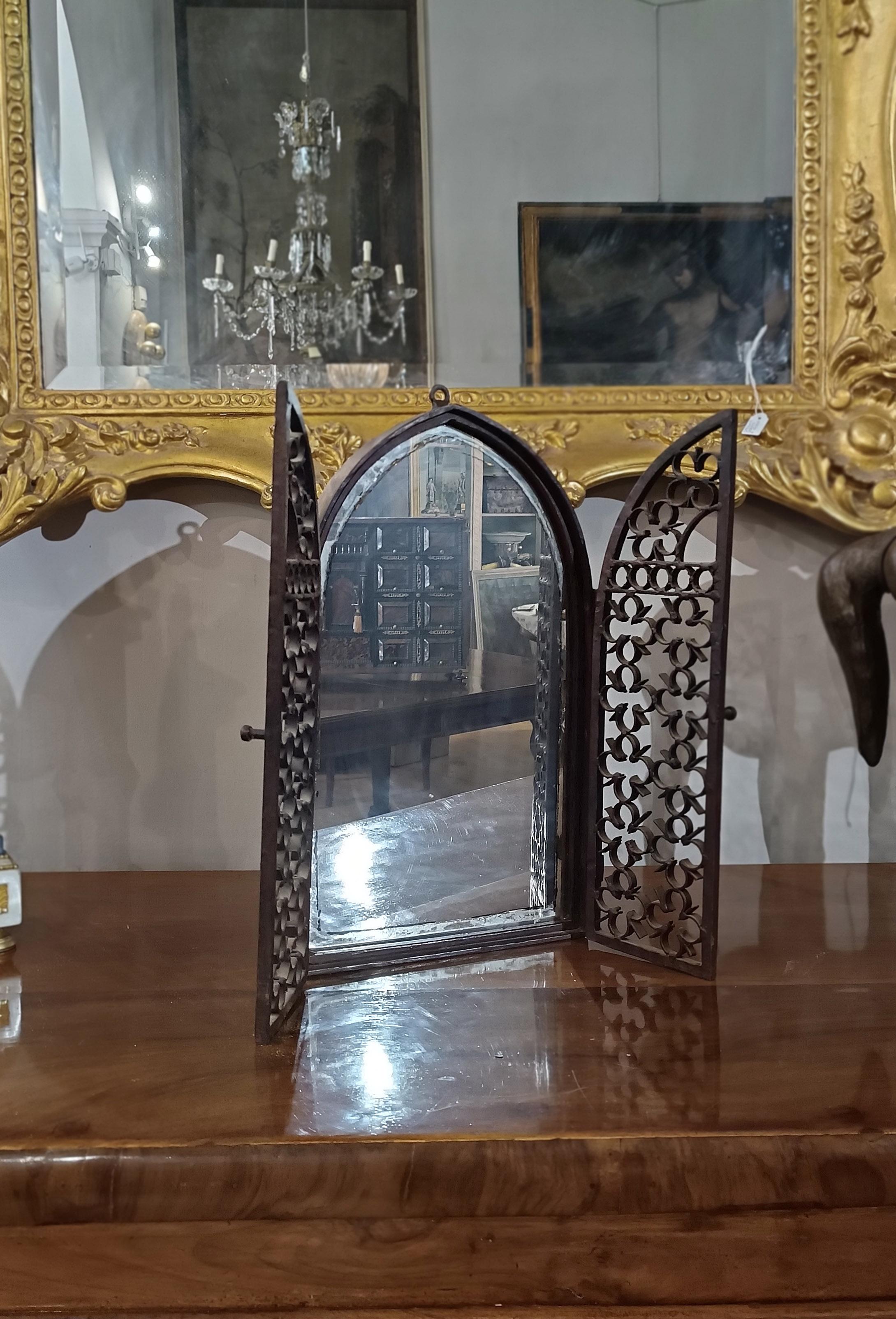 Elegant small mirror in wrought iron and hand-forged, in neo-Gothic style. Its “gate” shape evokes that of a Gothic window or stained glass window, characterized by an ogival profile. The two doors, decorated with floral and geometric motifs, can be
