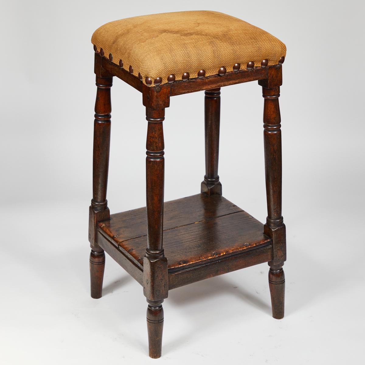 Tall wooden stool from 19th-century England with upholstered seat, brass stud detailing and lower shelf or footrest. The seat has been freshly reupholstered with cream-colored Belgian linen. Charmingly proportioned, this classic stool adds a