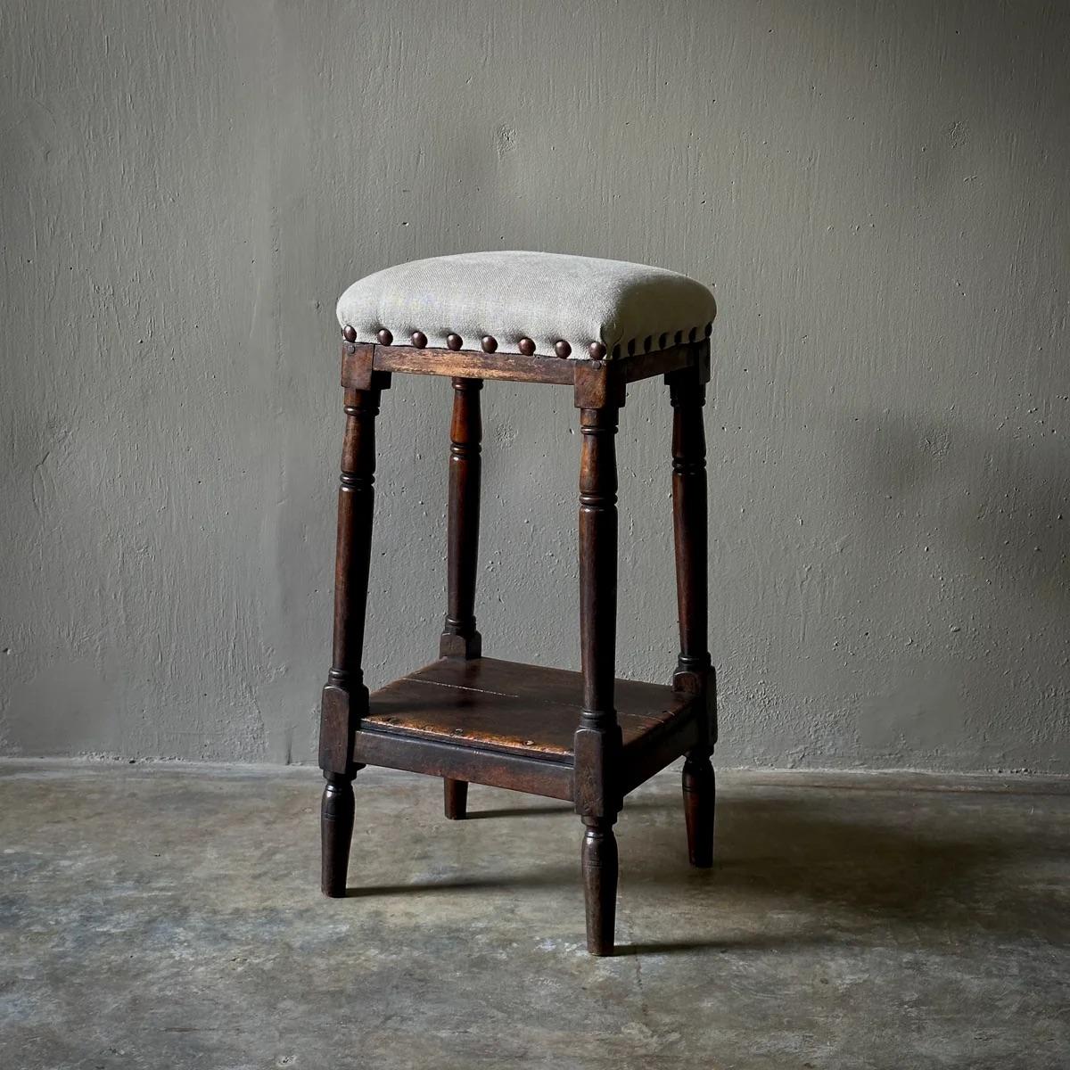 Questo sgabello inglese, realizzato in legno robusto, è dotato di un ripiano inferiore progettato con cura e di una seduta imbottita di recente in lino color crema, con borchie in ottone. La sua forma alta e slanciata la rende un'ottima scelta per i