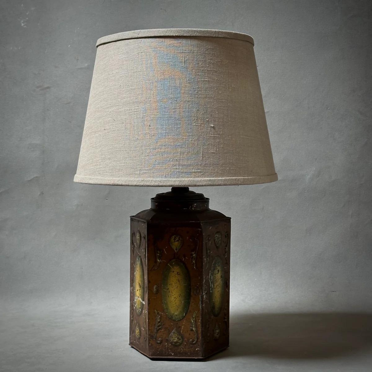 This English tole table lamp features deep rust and yellow hand-painted accents with a custom flax-toned linen shade. The hexagonal base is adorned with oblong discs on each panel, highlighted by subtle architectural flourishes. Its warm neutral