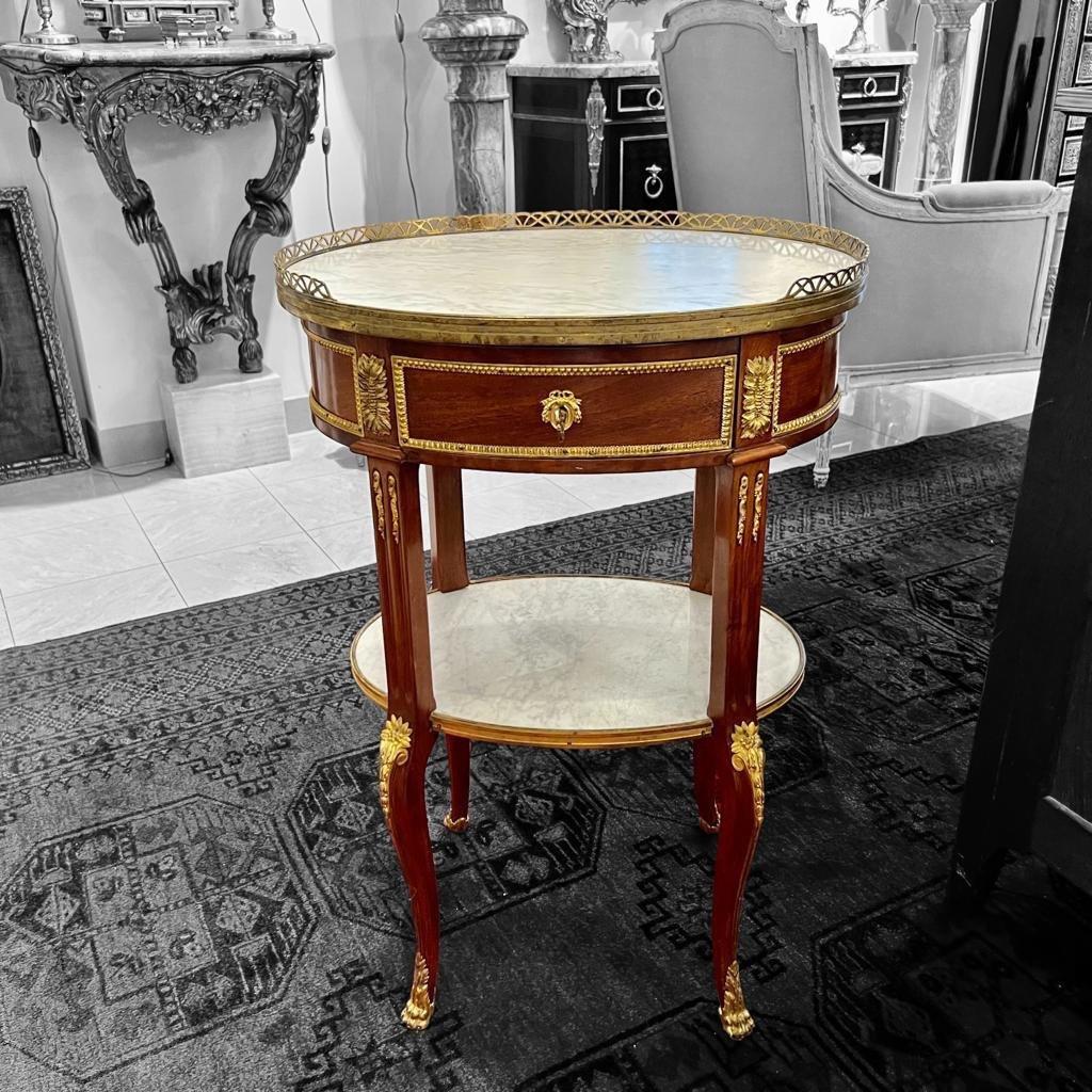 We present you this exquisite double-tiered Carrara marble gueridon in the Transition style of Louis XV. It is lavishly adorned with gilded bronze foliage ornamentation. The marble top is engirdled by a gilded bronze gallery and features a central