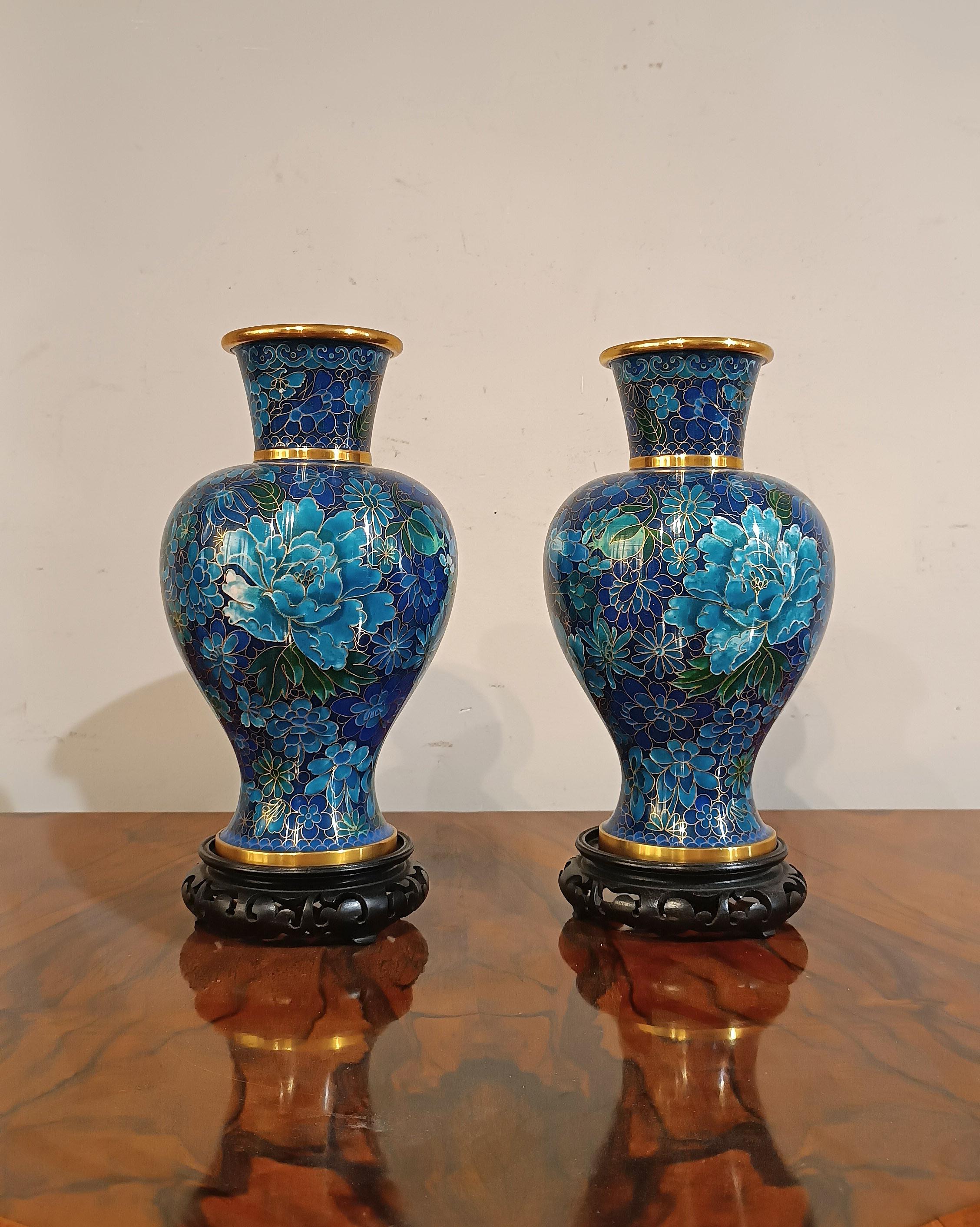 Beautiful pair of cloisonné vases in enameled and gilded metal, with hand-carved and ebonized wooden bases. The vases have a globular shape with a funnel neck. The enameled decorations on the body feature floral motifs, with fields of larger flowers