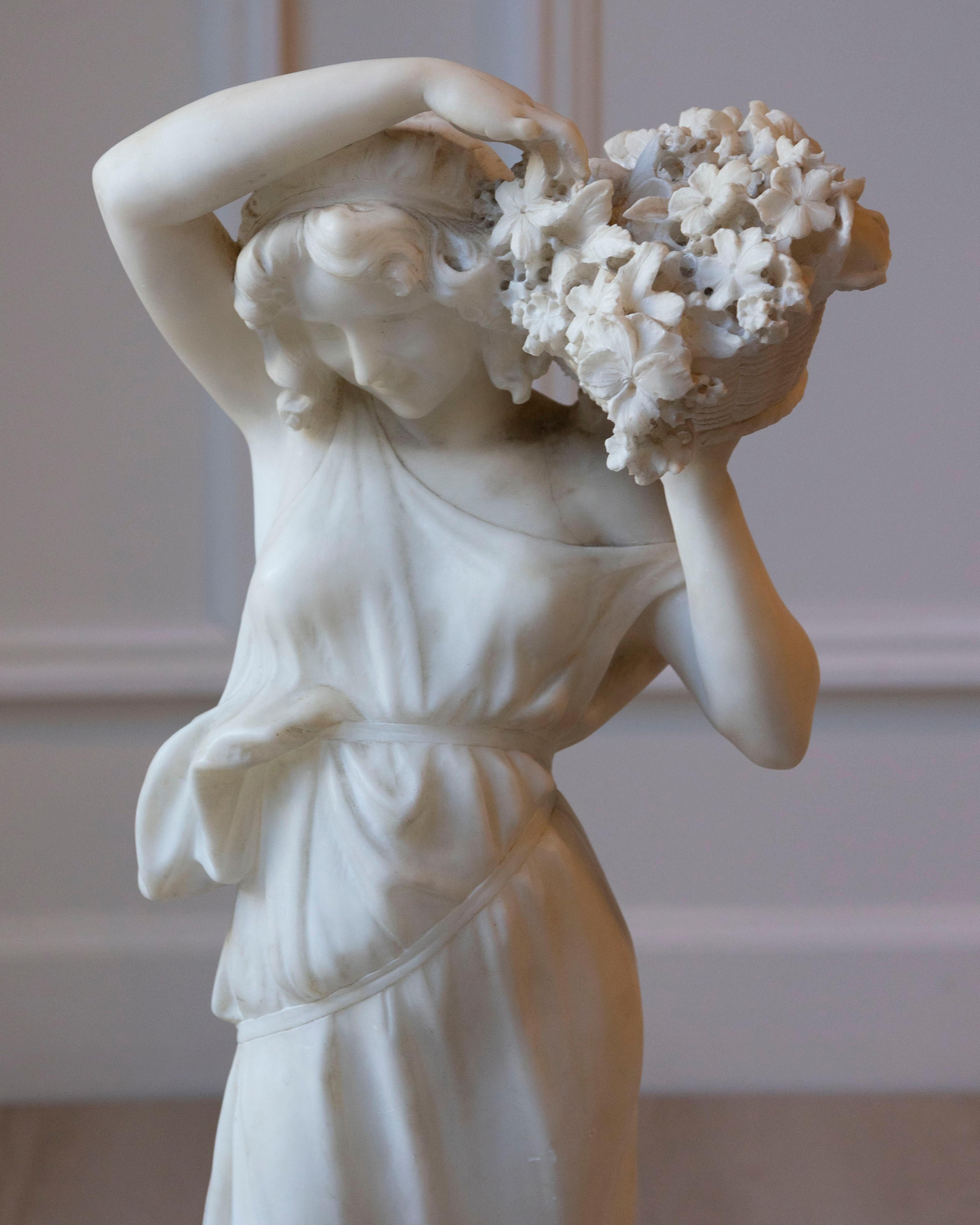 Una graziosa scultura in alabastro proveniente da Volterra, in Toscana, databile tra la fine del XIX e l'inizio del XX secolo, raffigurante una giovane fioraia che porta un cesto colmo di fiori. La figura è resa con morbidezza e naturale