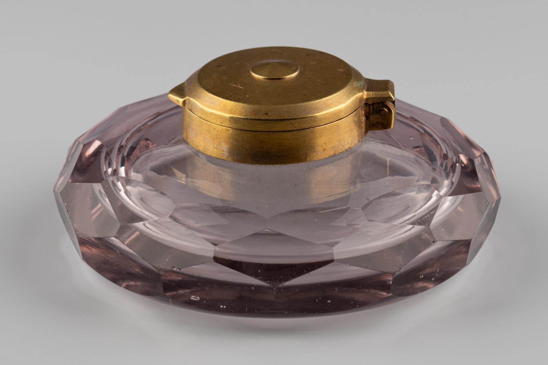 Tintero de cristal amatista facetado con tapa de latón

Origen: Francia
Material: vidrio, latón
Fecha: hacia 1900

Dimensiones:
A × P: aprox. 6 × 12 cm

Descripción:
Tintero bajo y circular con cuerpo facetado de cristal ligeramente tintado. El