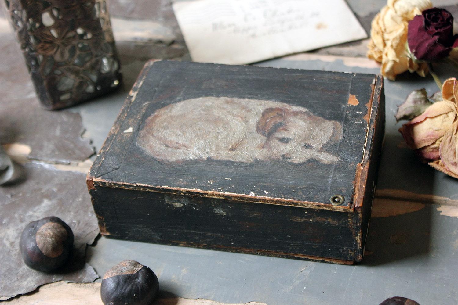 The rectangular black painted cigar box having a pin fastened lid revealing a vacant interior, the front painted with a naïve study of a curled-up border terrier and surviving from late Victorian period England.

This box has obvious signs of use