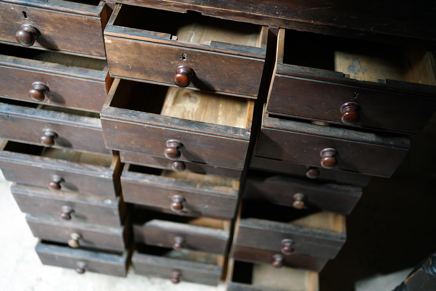 In good original condition, the stained pine flight or bank of twenty-one drawers, each with knob handles, having seven rows of three drawers, consisting of four smaller rows graduating to three deeper rows each with grooved borders, the whole