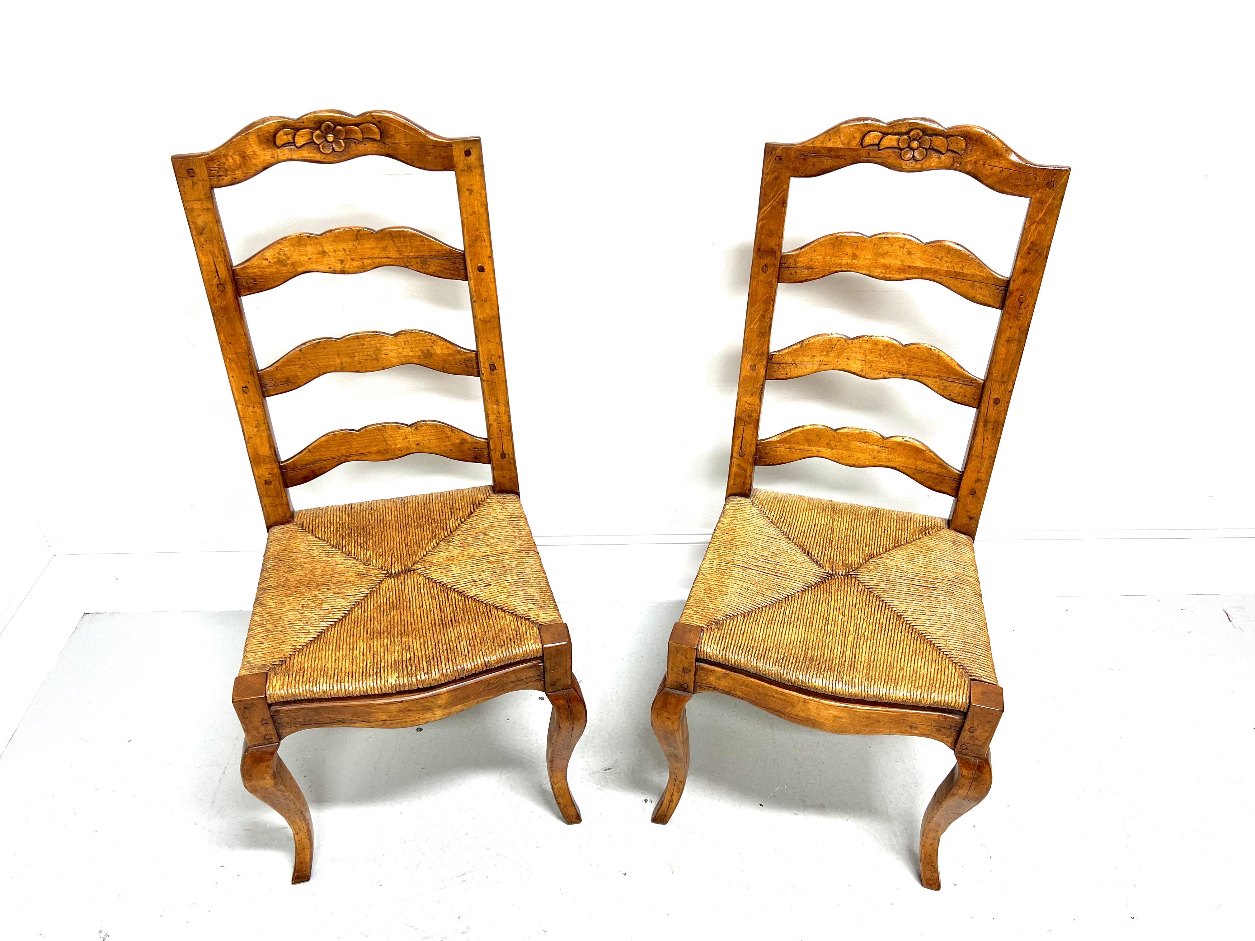 A pair of French Country style dining side chairs, unbranded. Solid nutwood with a distressed finish, ladder back design, decoratively carved crest rail, rush seats, carved apron, and curved legs. Made in the USA, in the late 20th