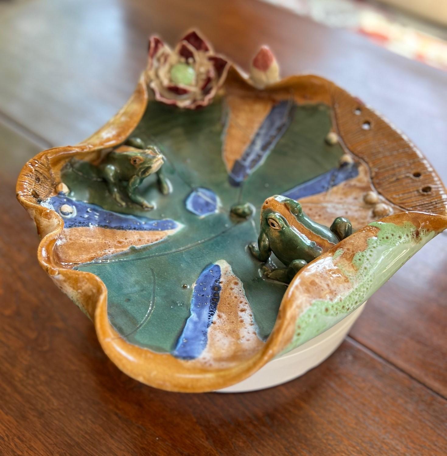 Late 20th C. a hand-crafted ceramic decorative bowl. The design prominently features two small frogs sitting on a large, stylized lily pad, with delicate details such as a flower and additional buds. The frogs and the lily pad are hand-sculpted. The