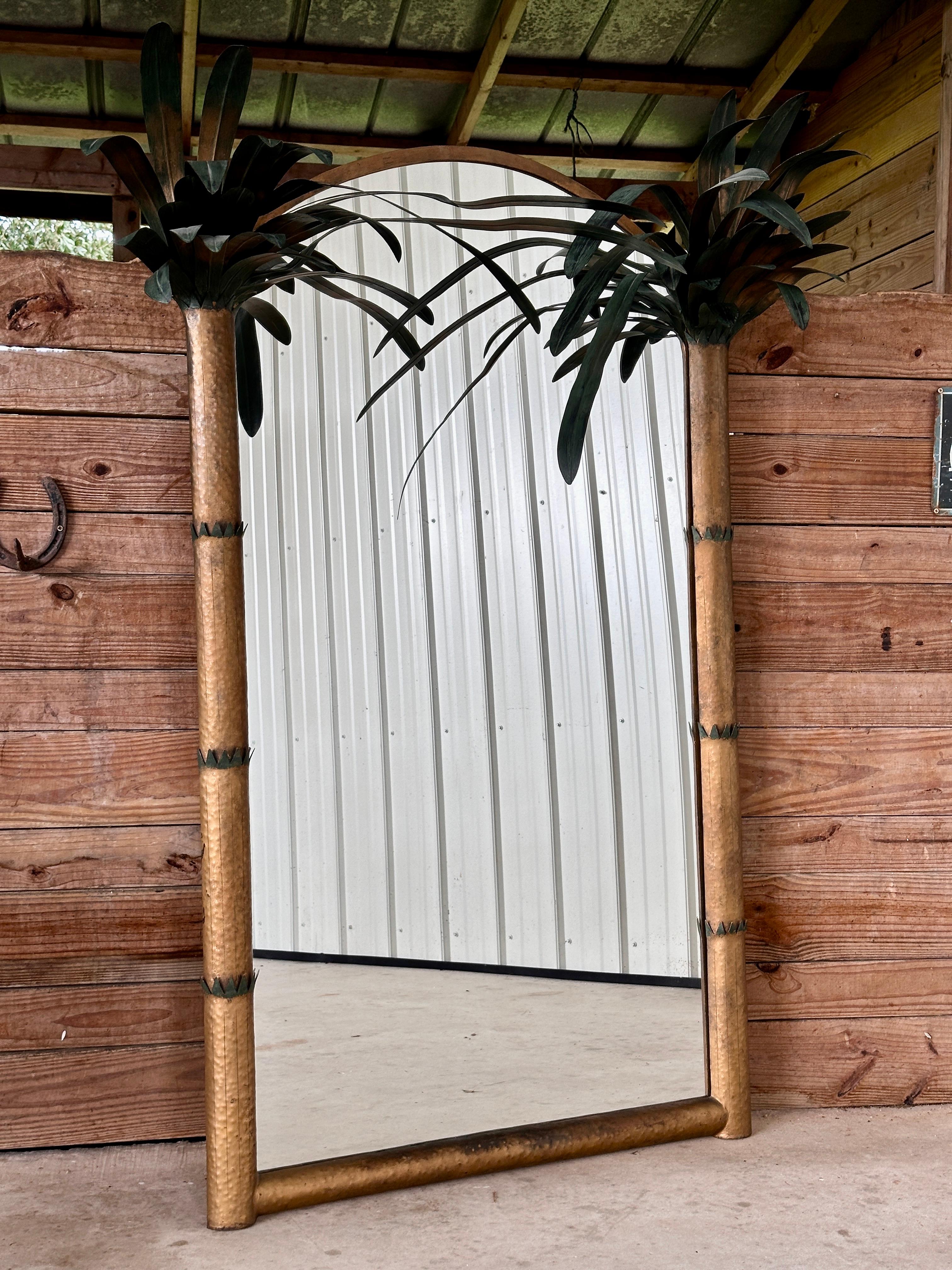 This striking vintage wall mirror features a sculptural design with hand-painted metal tole palm trees, bringing an exotic and tropical flair to any space.
The arched mirror is framed by two textured faux bamboo columns that extend upwards into lush