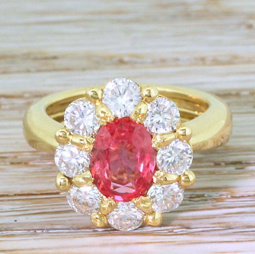 Padparadscha sapphires are as scarce as they are beautiful. This stunning example displays a bright, vivid orangey pink, the colour of which is emphasised by the eight high white and clean round brilliant cut diamonds in the surround. The open