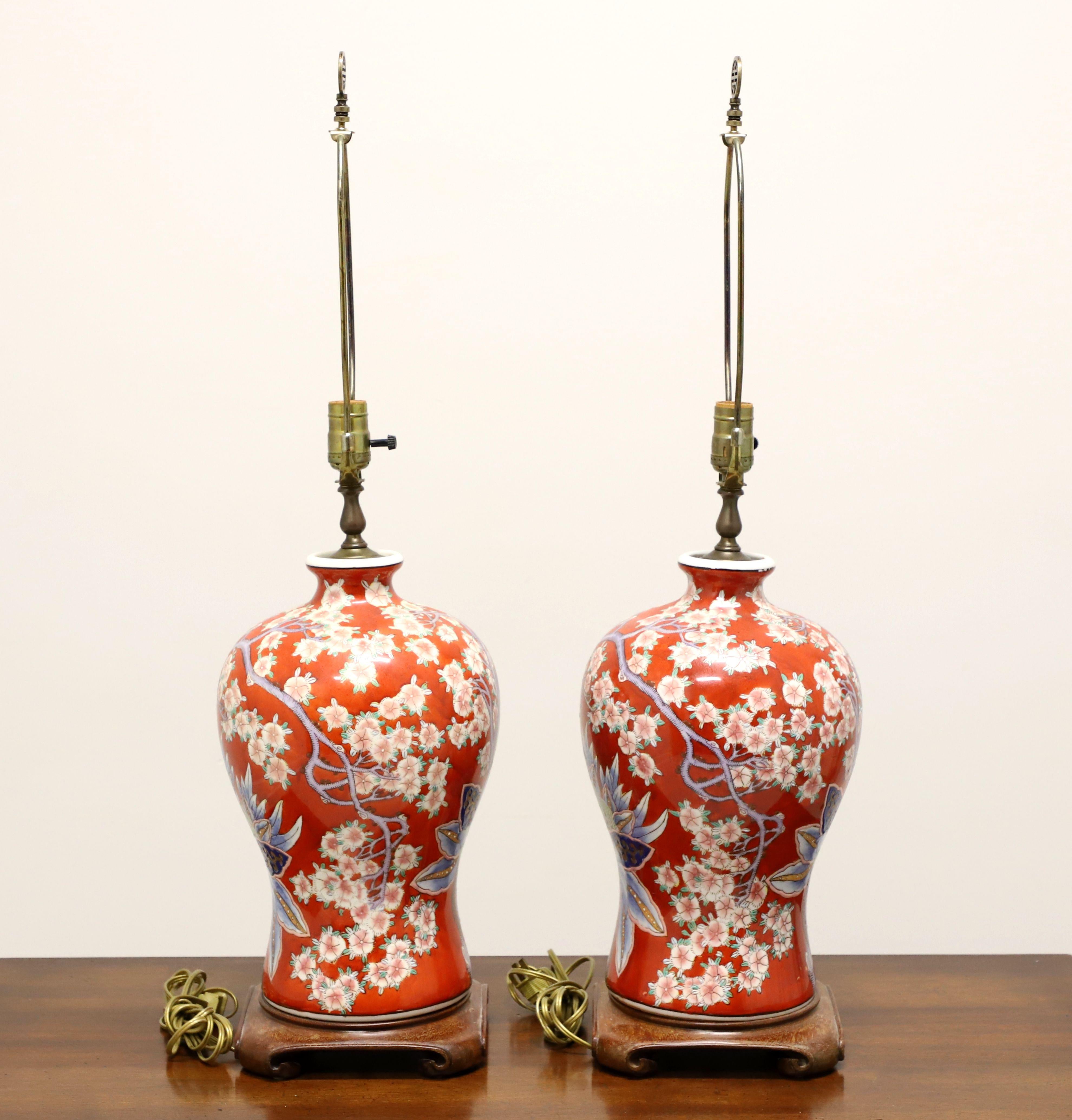 A pair of Asian Chinoiserie style table lamps, unbranded. Made of porcelain in an urn style, hand painted, with a red base color, pink & white cherry blossoms, blue, green & brown floral accents, and on a carved wood base. Has a metal harp and
