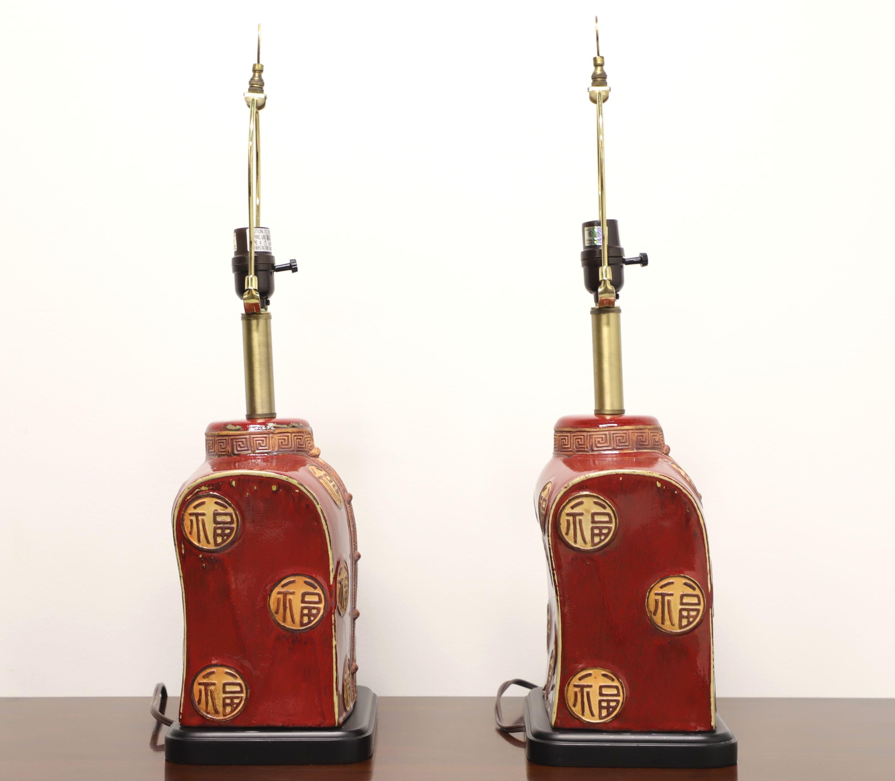 A pair of Asian style table lamps, unbranded. Made of porcelain to represent a Chinese Tang Suit in red and gold, on a black painted wood base. Has bass neck, harps and finials. Single standard bulb socket and 3-way rotary switch. Likely made in