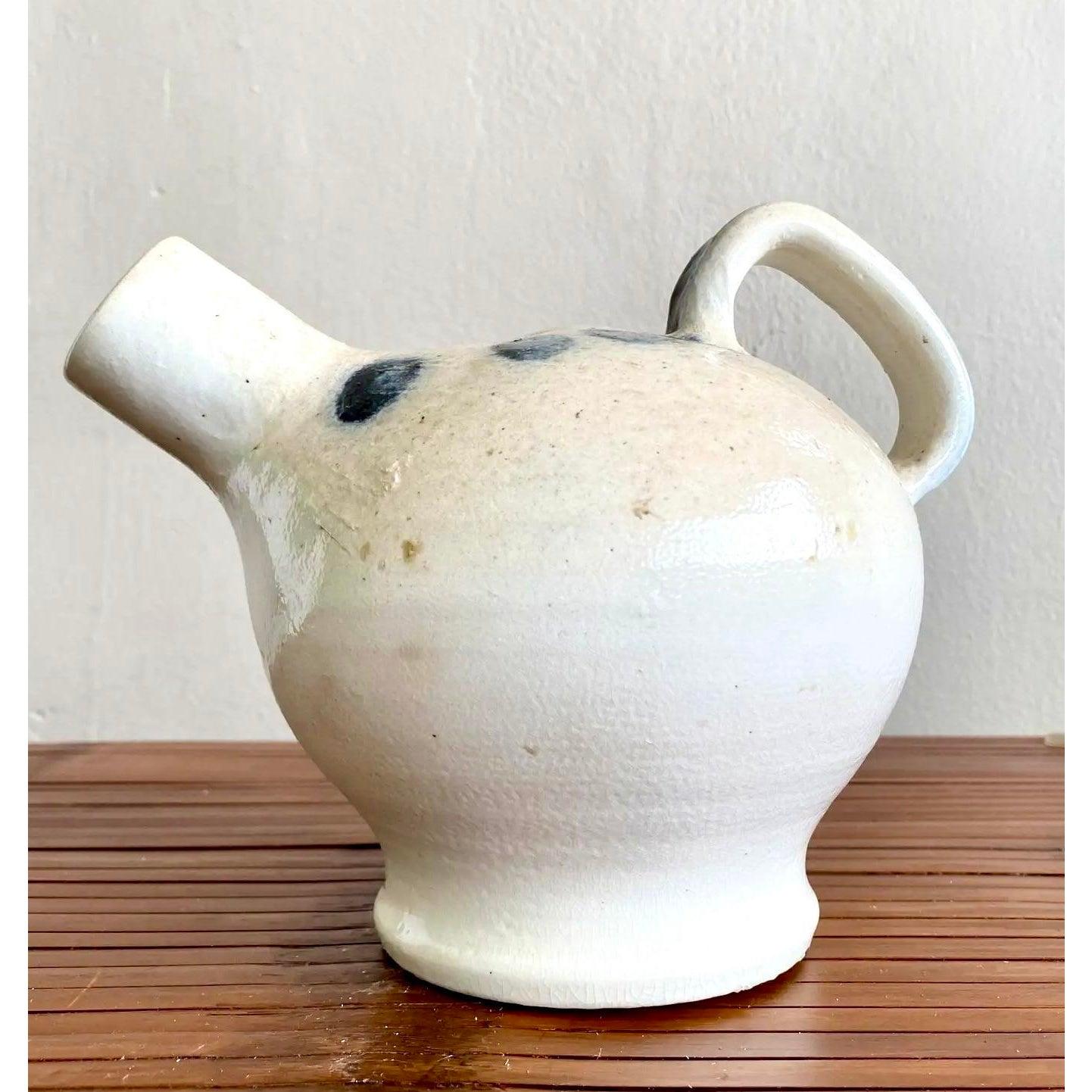 Eine charmante, handgefertigte Studiotöpferei mit einer runden Form, einem kurzen Ausguss und einem Henkel mit Schleife. Die cremefarbene Glasur wird von malerischen blauen Punkten akzentuiert, die ihr eine rustikale und doch verspielte Ausstrahlung