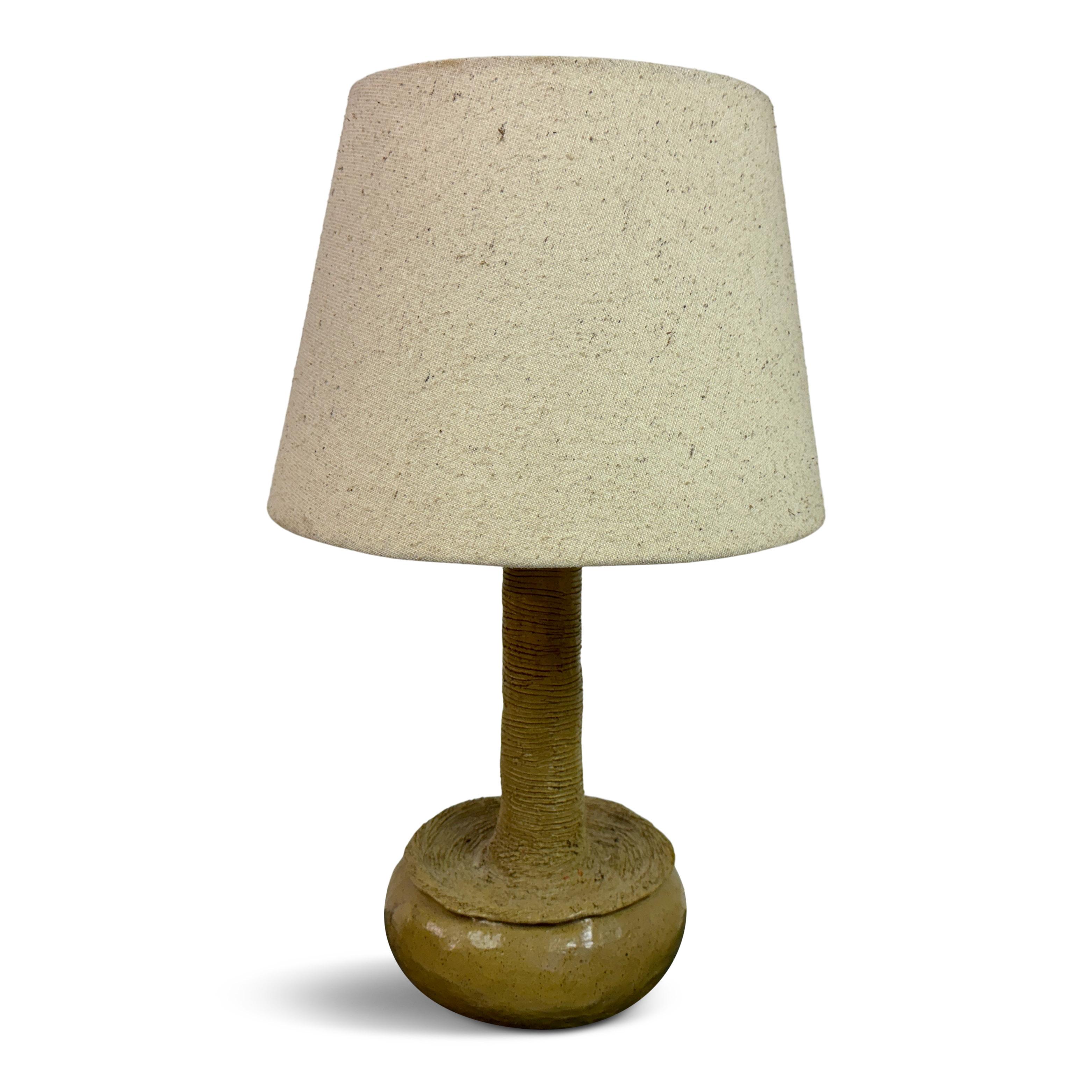 This is a striking brutalist ceramic lamp, featuring a unique and earthy design. The base is a bulbous, textured ceramic form with a natural, rough finish that emphasizes the raw beauty of the material. Rising from the base is a cylindrical neck,