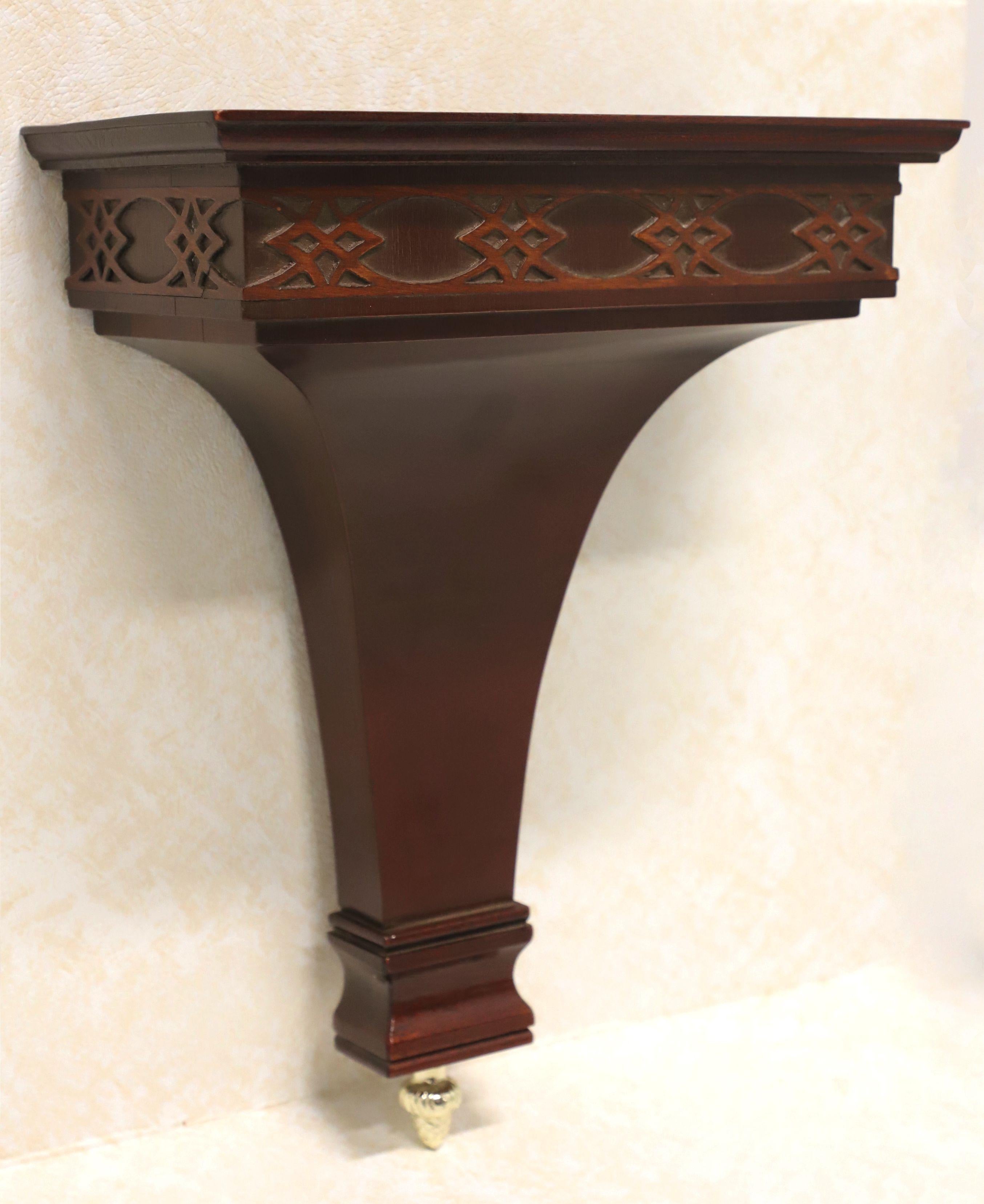 A Chippendale style wall bracket shelf, unbranded. Solid mahogany with flat display surface, decorative fretwork to apron, and brass finial at bottom. Dual brass screw holders on back for wall hanging. Made in the USA, in the late 20th