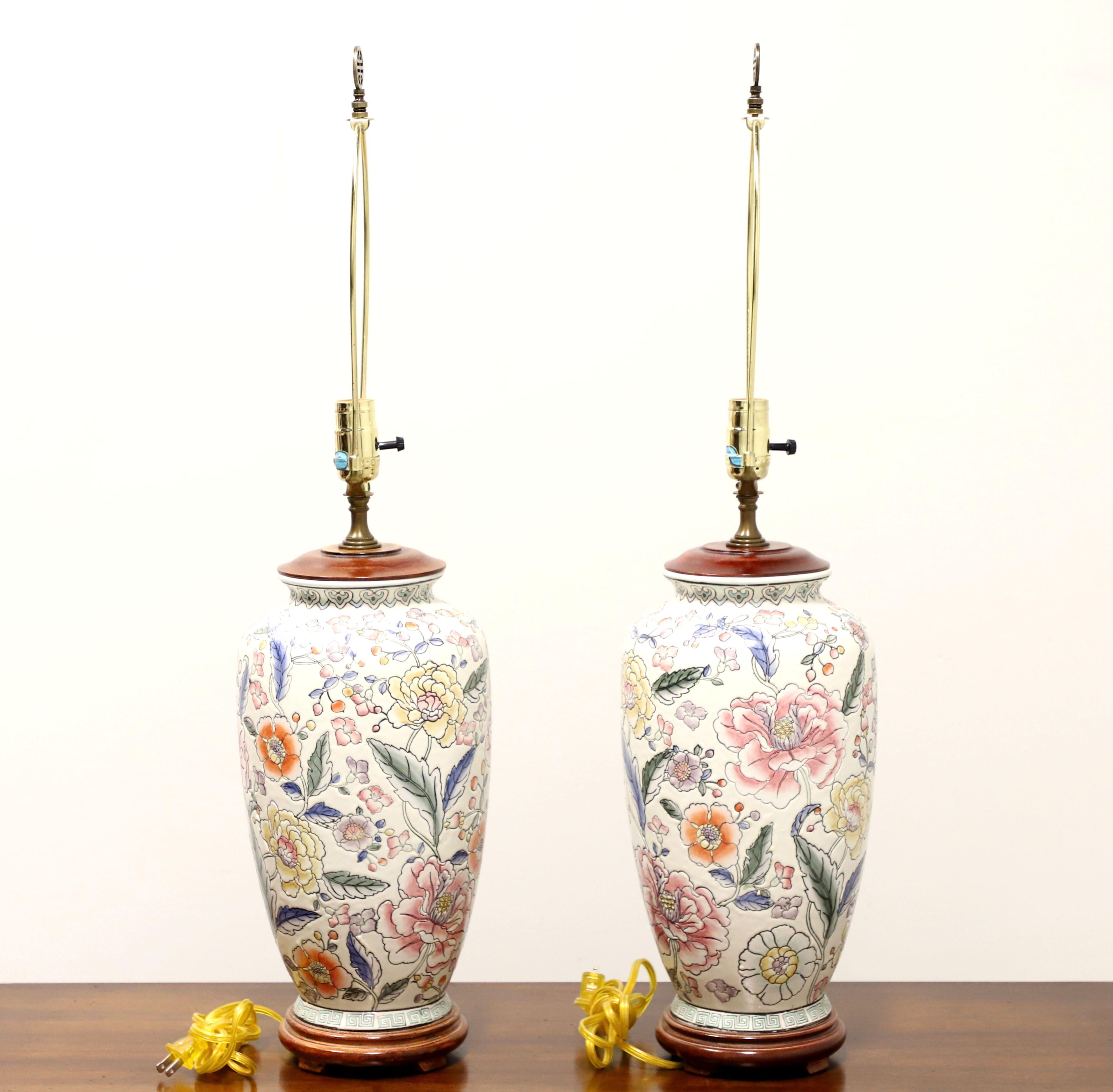 A pair of Asian Chinoiserie style table lamps, unbranded. Made of porcelain in an urn style, hand painted, with a creamy white base color, pink, yellow, orange, blue & green floral motif, a wood lid to top, and on a carved wood base. Has a metal