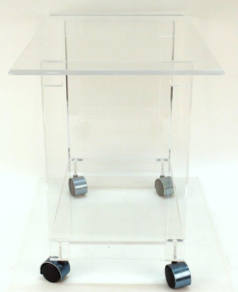 A clear Lucite bar cart from the late 20th century on casters with two shelves. The cart primarily consists clear Lucite, which gives the bar-cart a 