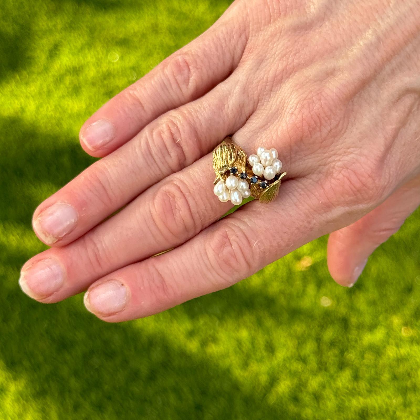 This late-20th century vintage tulip ring features a stunning combination of cultured pearls, sapphires, and 14K yellow gold. The design showcases two bypass tulips, each with petals made of lustrous cultured pearls, and blue sapphire accents placed