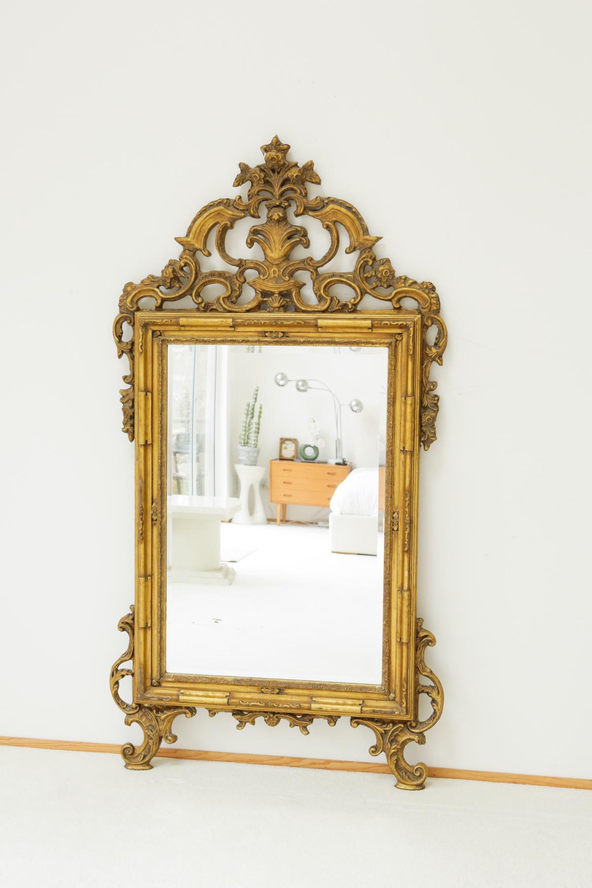 A vintage Louis XVI style mirror created from giltwood, fit for royalty. This grand and uniquely shaped mirror features elaborate carvings of various floral and foliate motifs. There is an emphasis on visual lightness and harmony, much of its