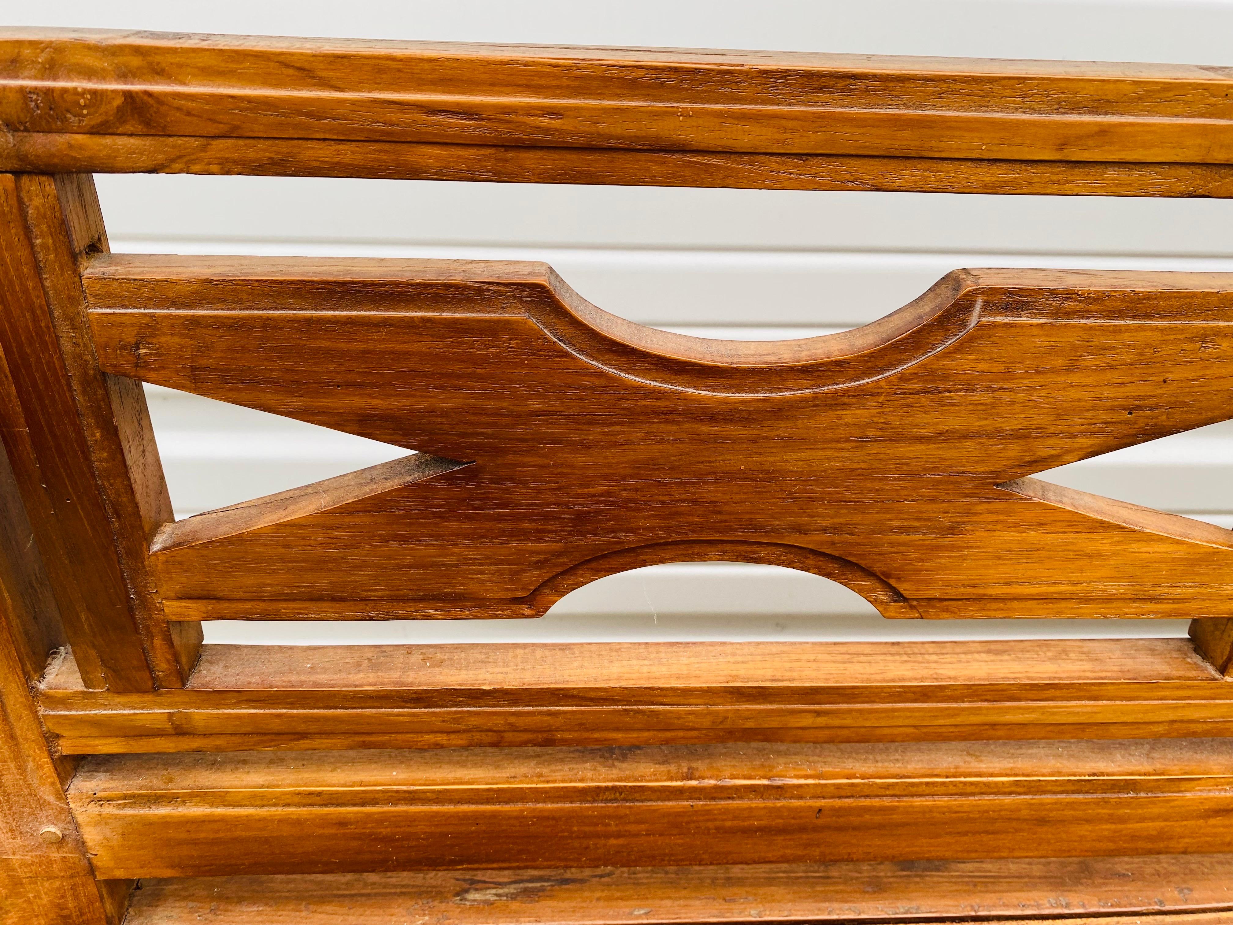 Il s'agit d'un banc de décoration en teck fabriqué à la main à la fin du 20e siècle. Ce banc est doté d'un dossier en X sculpté à la main et d'un accoudoir aux courbes douces. Ce long banc présente une finition très polie de couleur ambre. Ce banc a