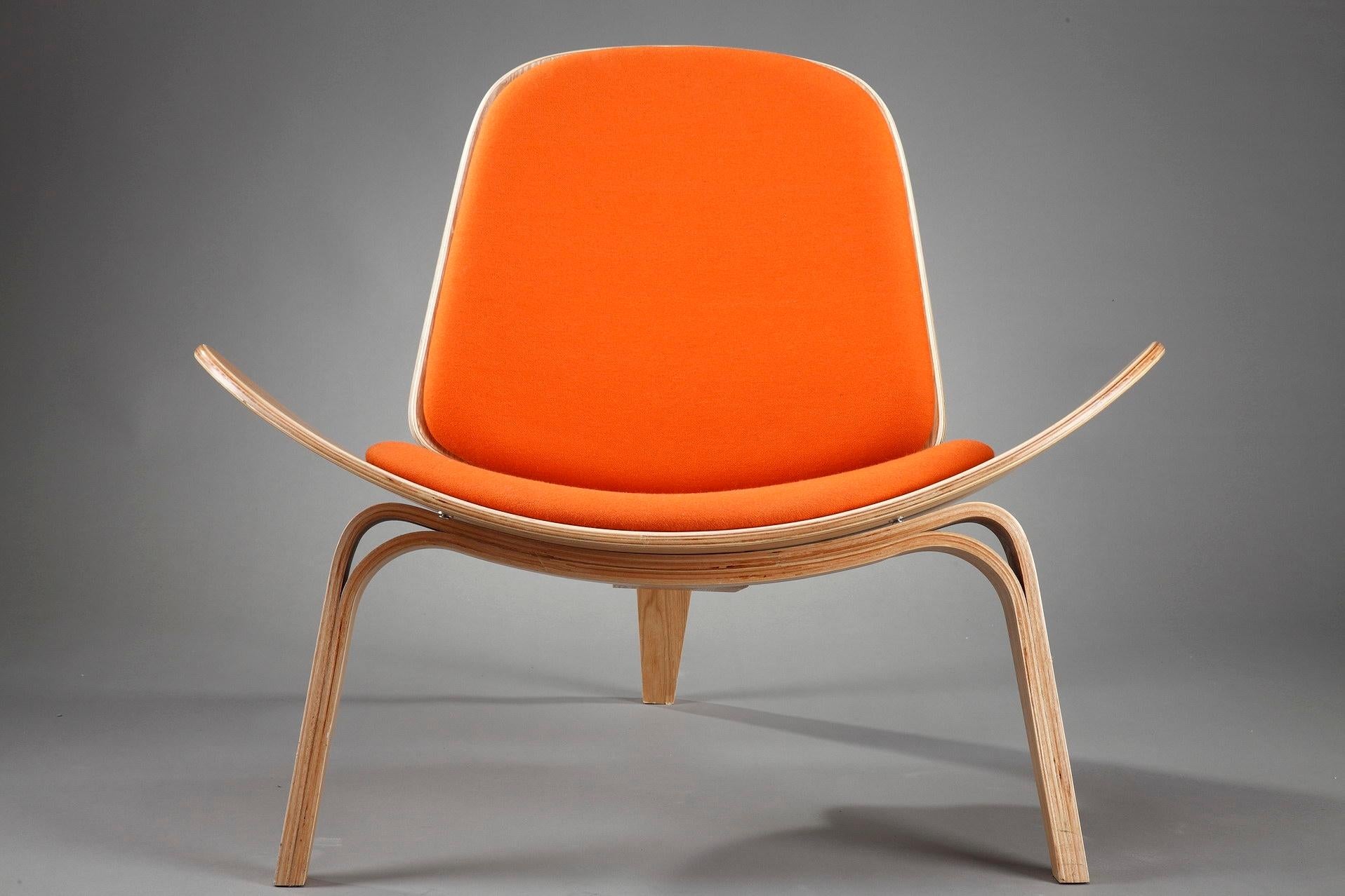 The CH07 shell chair was created in 1963 by the Danish designer Hans J. Wegner (1914-2007). It is crafted in bent plywood with orange fabric upholstery, and rests on three legs. The shell chair achieves a floating lightness due to its wing-like seat