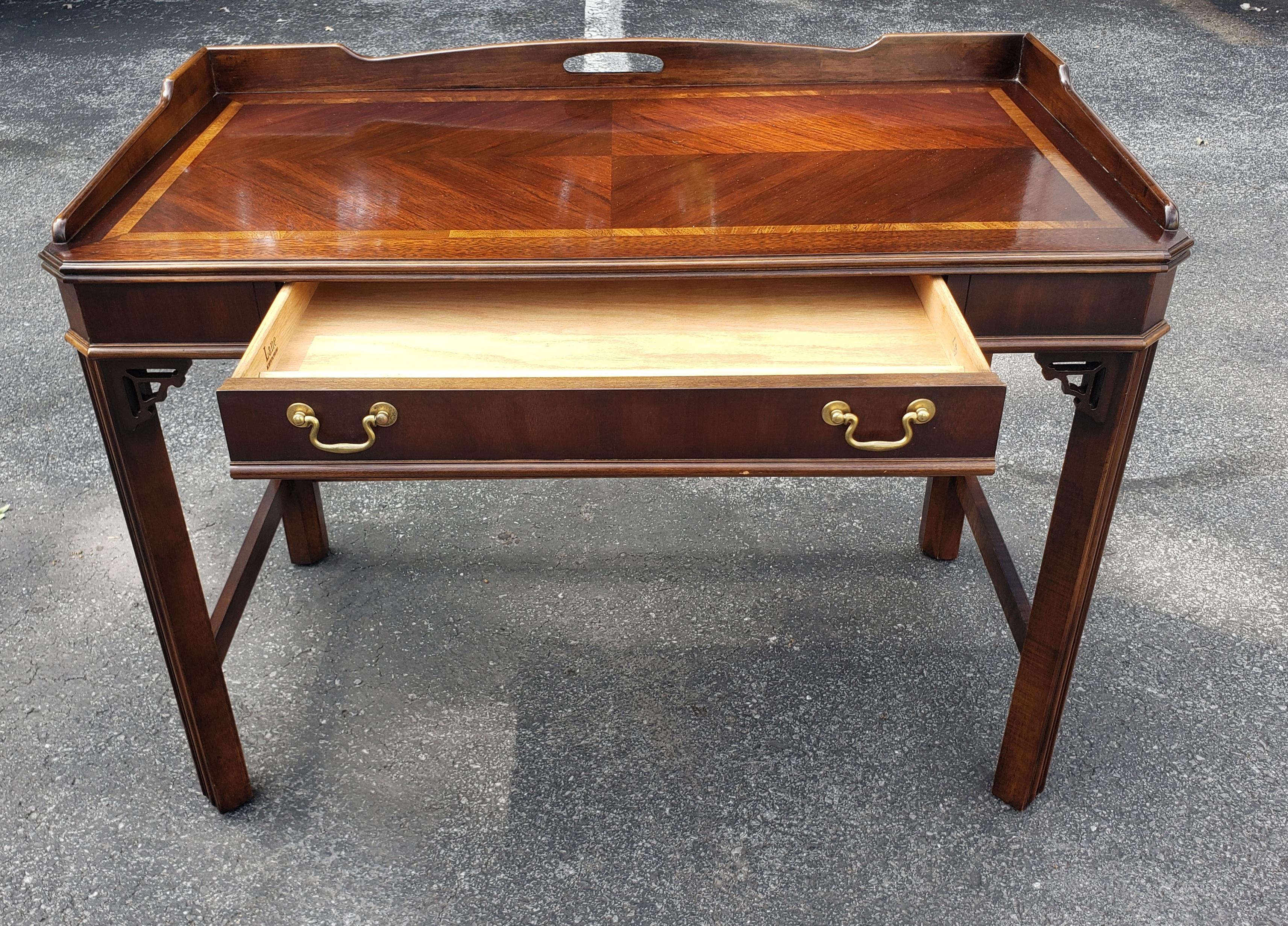 This is a charming little vintage Lane Furniture Chinese Chippendale, Mahogany wood writing desk. This features a beautiful banded inlaid center piping and a stretcher base, with brass handles on either side. It has a center drawer and fretwork