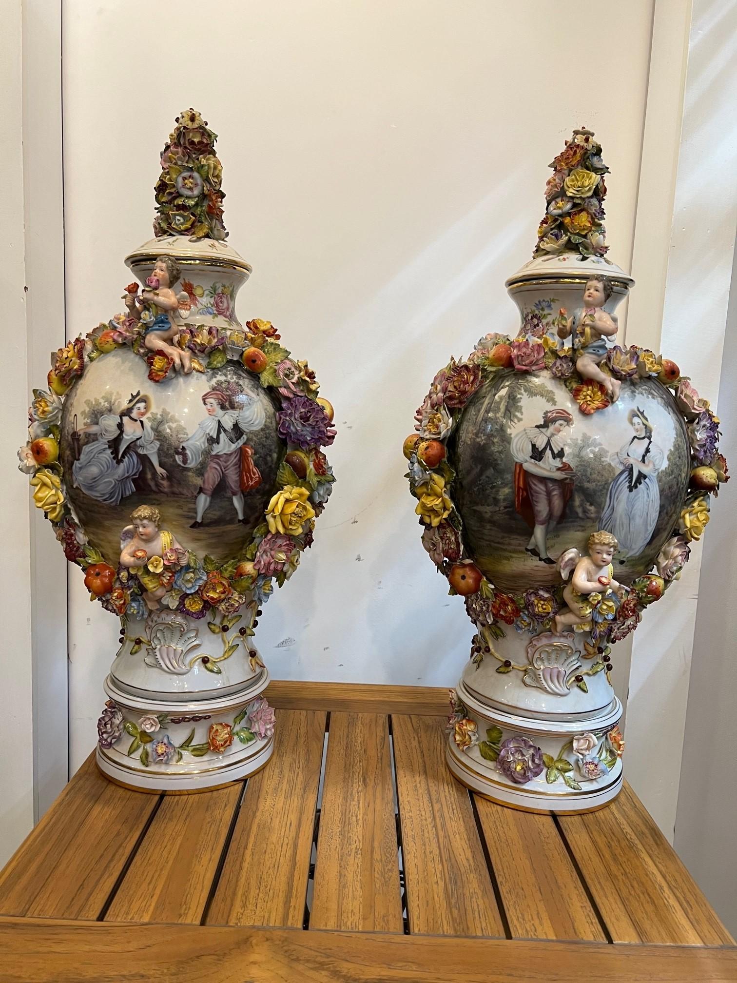 A beautiful pair of Dresden style hand painted porcelain urns with lids. A very impressive and decorative pair of Dresden style lidded porcelain urns. Classical romantic hand painted scenes with  pierced lids on a raised round pedestal base. The