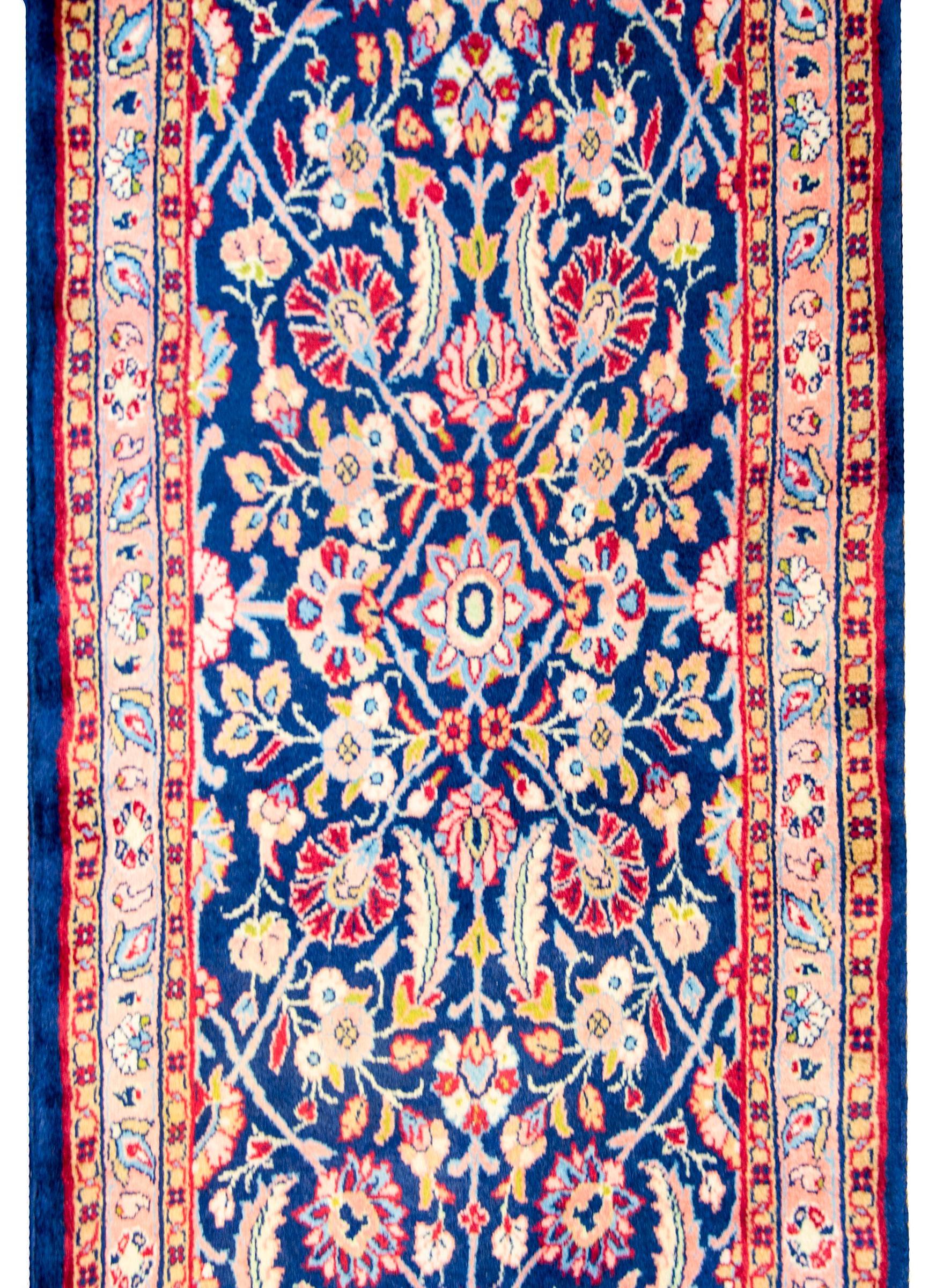 A beautiful late 20th century Persian lilihan runner with three floral medallions amidst a field of myriad flowers and scrolling vines woven in light indigo, pink, green, gold, cream, and dark cranberry, all on a dark indigo field. The border is