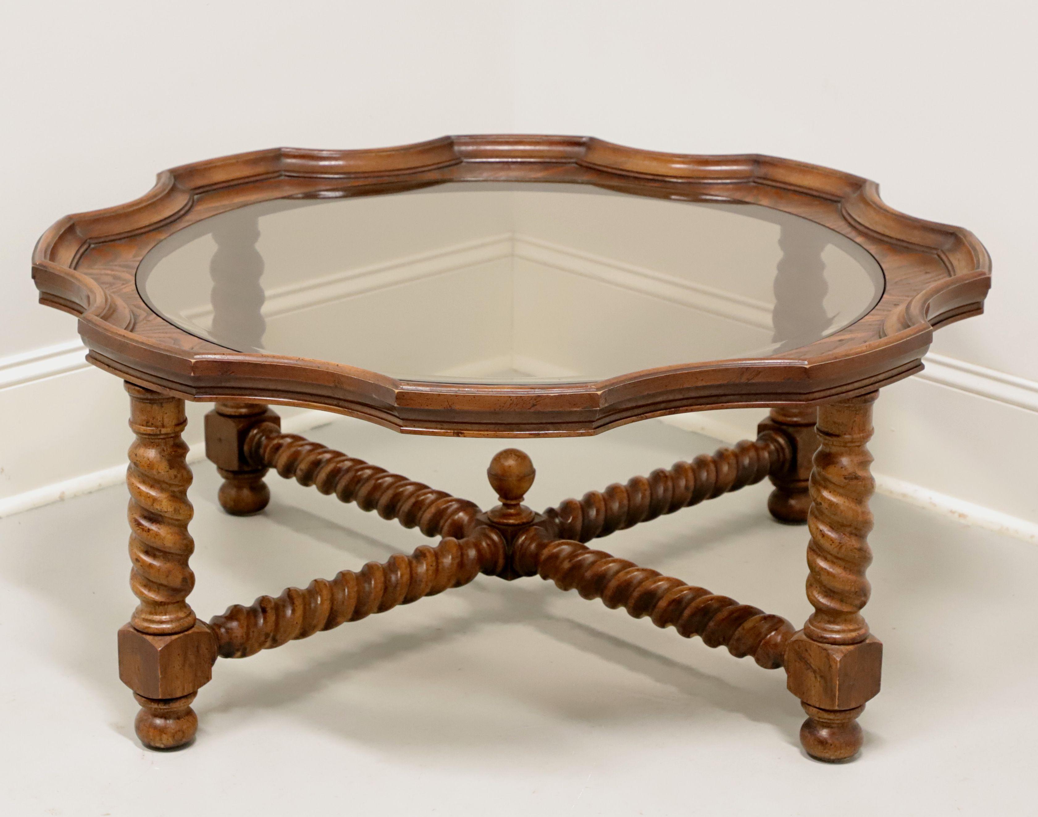 A Jacobean style round glass top coffee table, unbranded, similar in quality to Baker. Solid oak with tiger oak inlay to edge of scalloped top, inset ogee edge smoked glass, “X” shaped barley twist stretcher with center finial, and barley twist legs