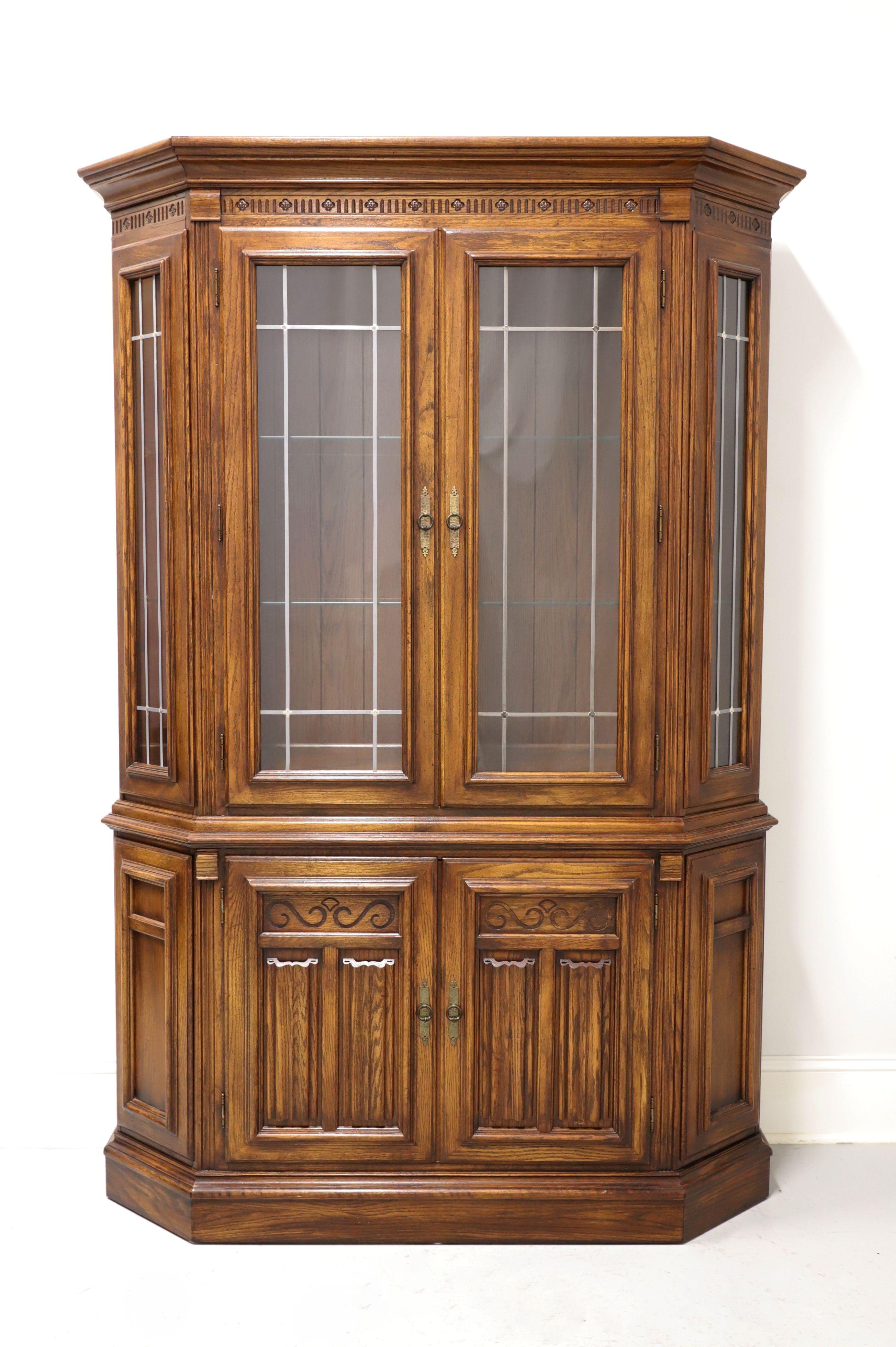 A Jacobean style china cabinet, unbranded, similar quality to Thomasville. Oak with crown & dentil molding to the top, brass hardware, and a solid base. Upper cabinet has two metal paned glass doors & two angled side panels, is lighted, and has two