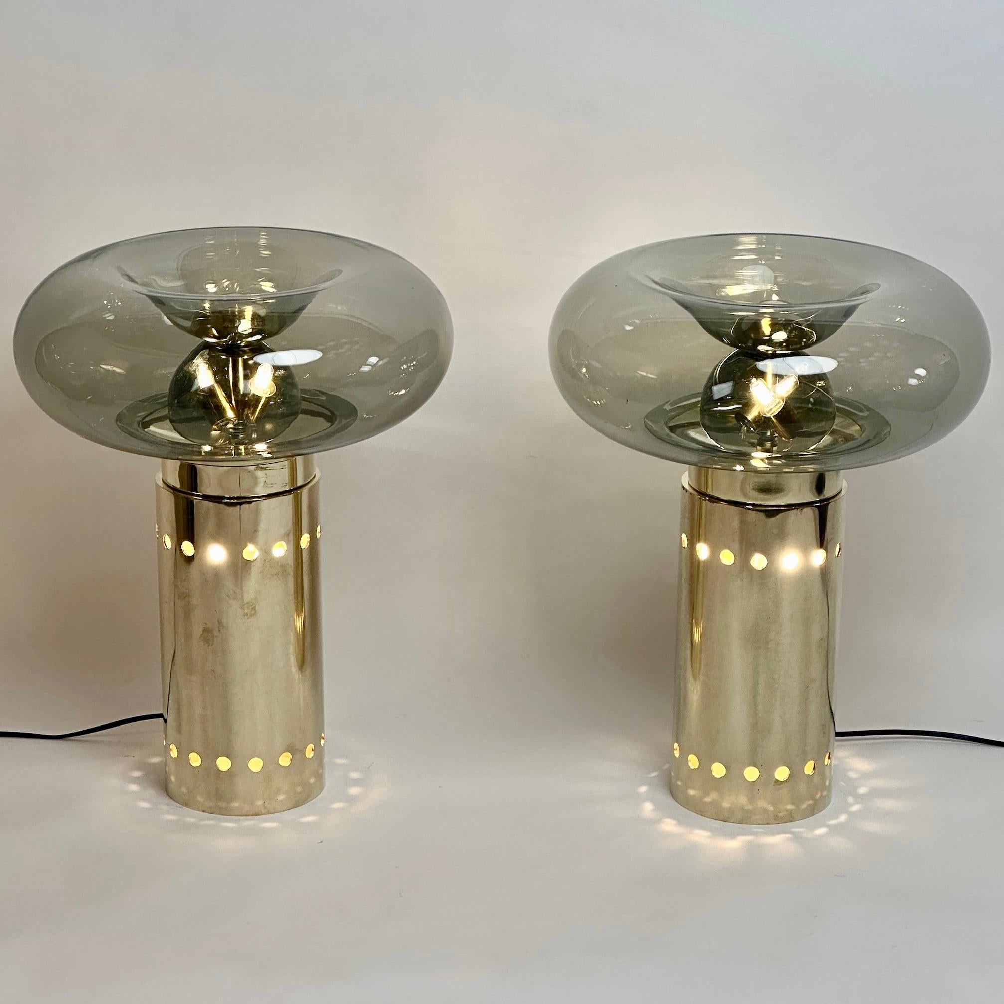 This pair of modern table lamps with smoked grey blown Murano art glass diffusers fit almost any kind of environment and they are perfect to be placed either on any leaving room or any bedroom.
The circular brass basement resembling a smooth column