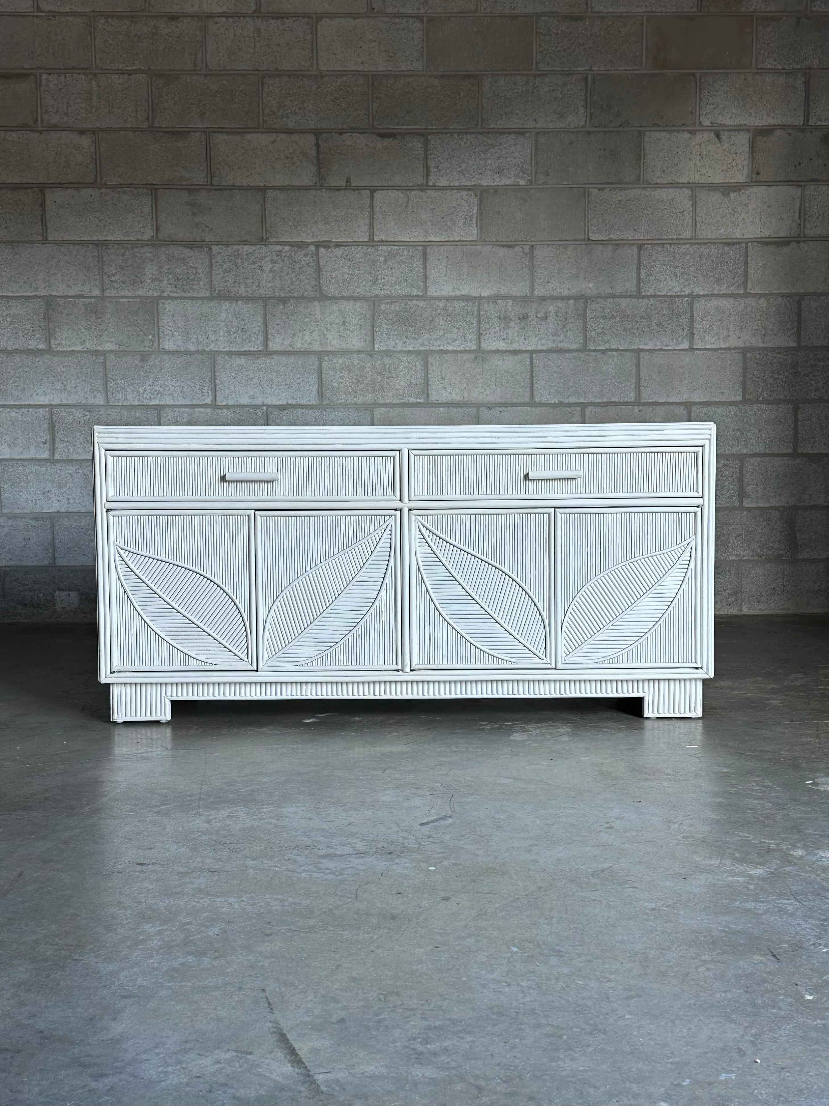 A stunning pencil reed sideboard in original white paint. Features two large compartments accessed by a pair of doors on either side. Above each compartment is a drawer. Door fronts feature a leaf pattern.