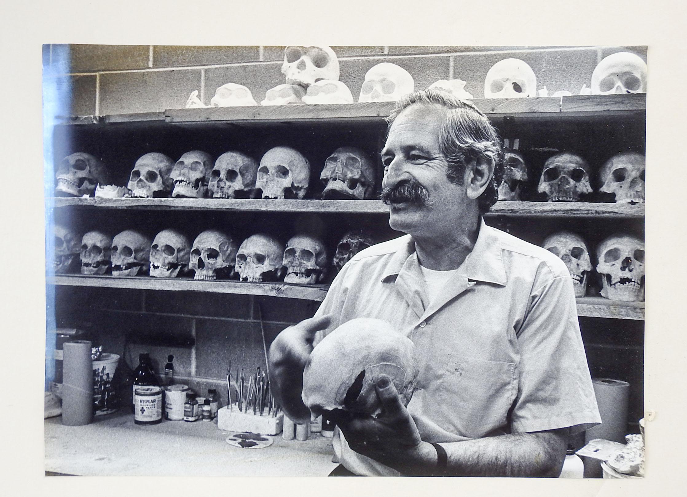 Late 20th century black and white photograph of a man and shelves of human skulls. Likely a university or museum. Unsigned. Unframed.